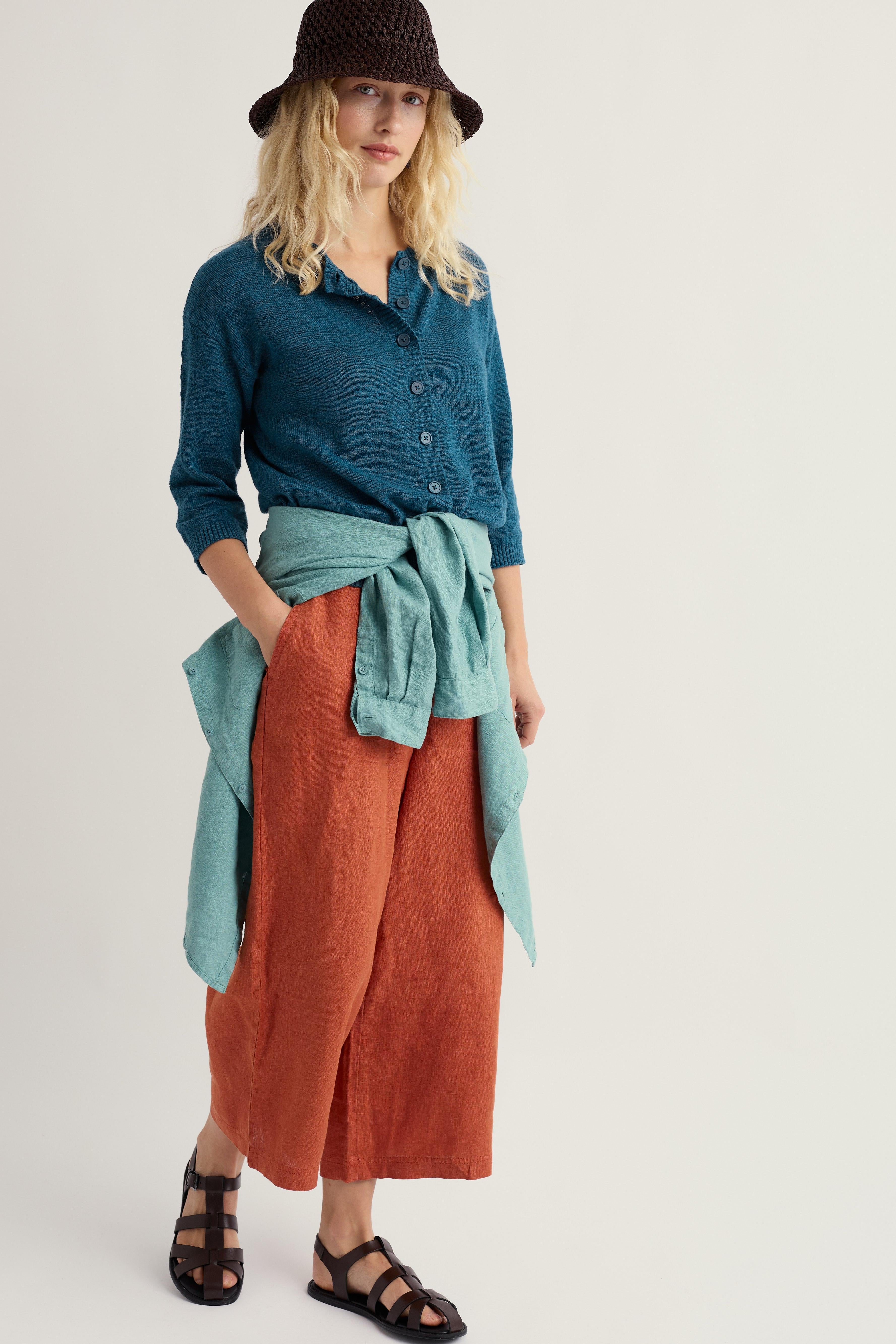 Woman wearing a teal organic cotton cardigan, rust-colored pants, and a black hat on a white background