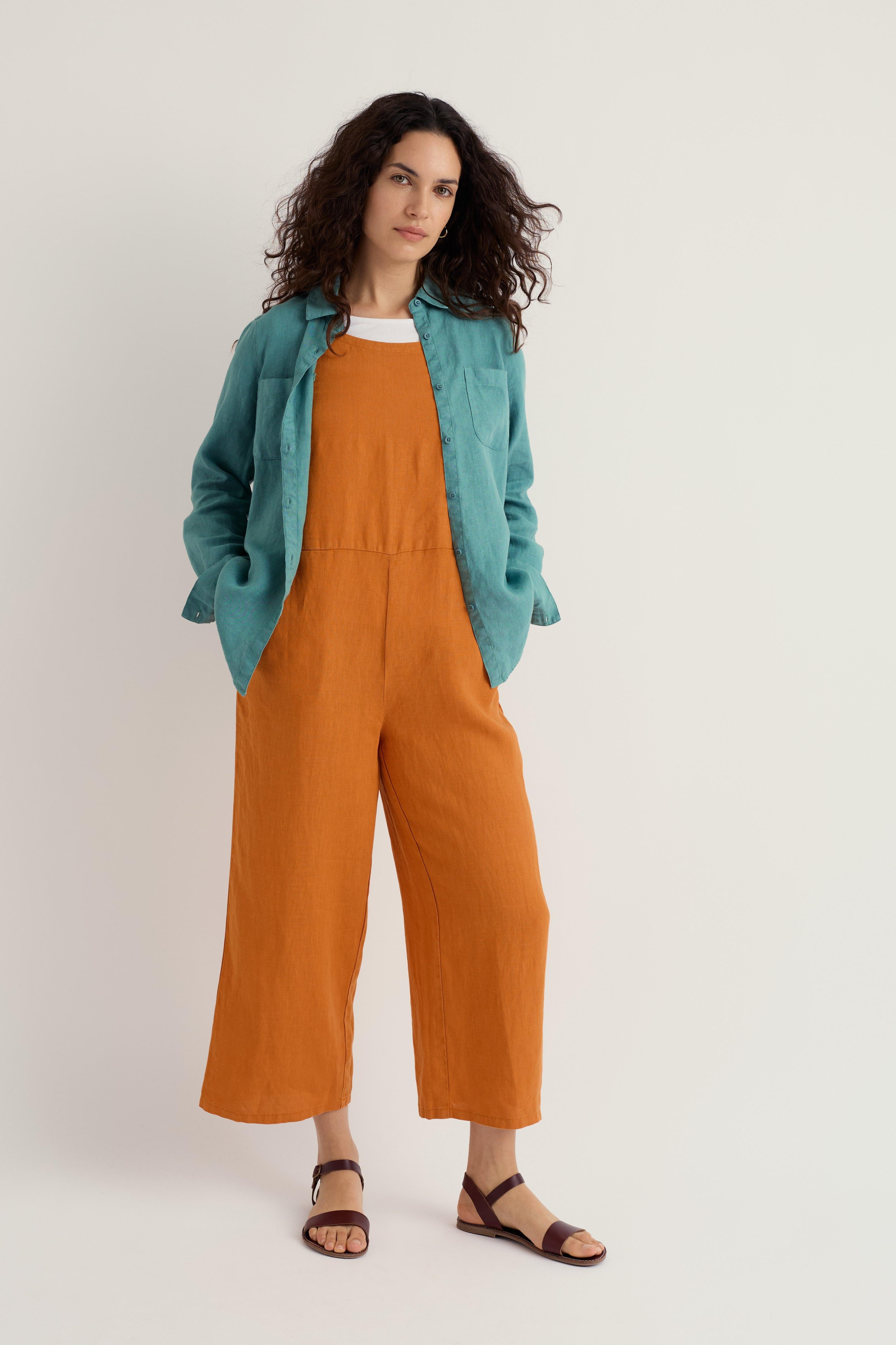 Woman wearing an orange linen jumpsuit with a teal jacket on a white background