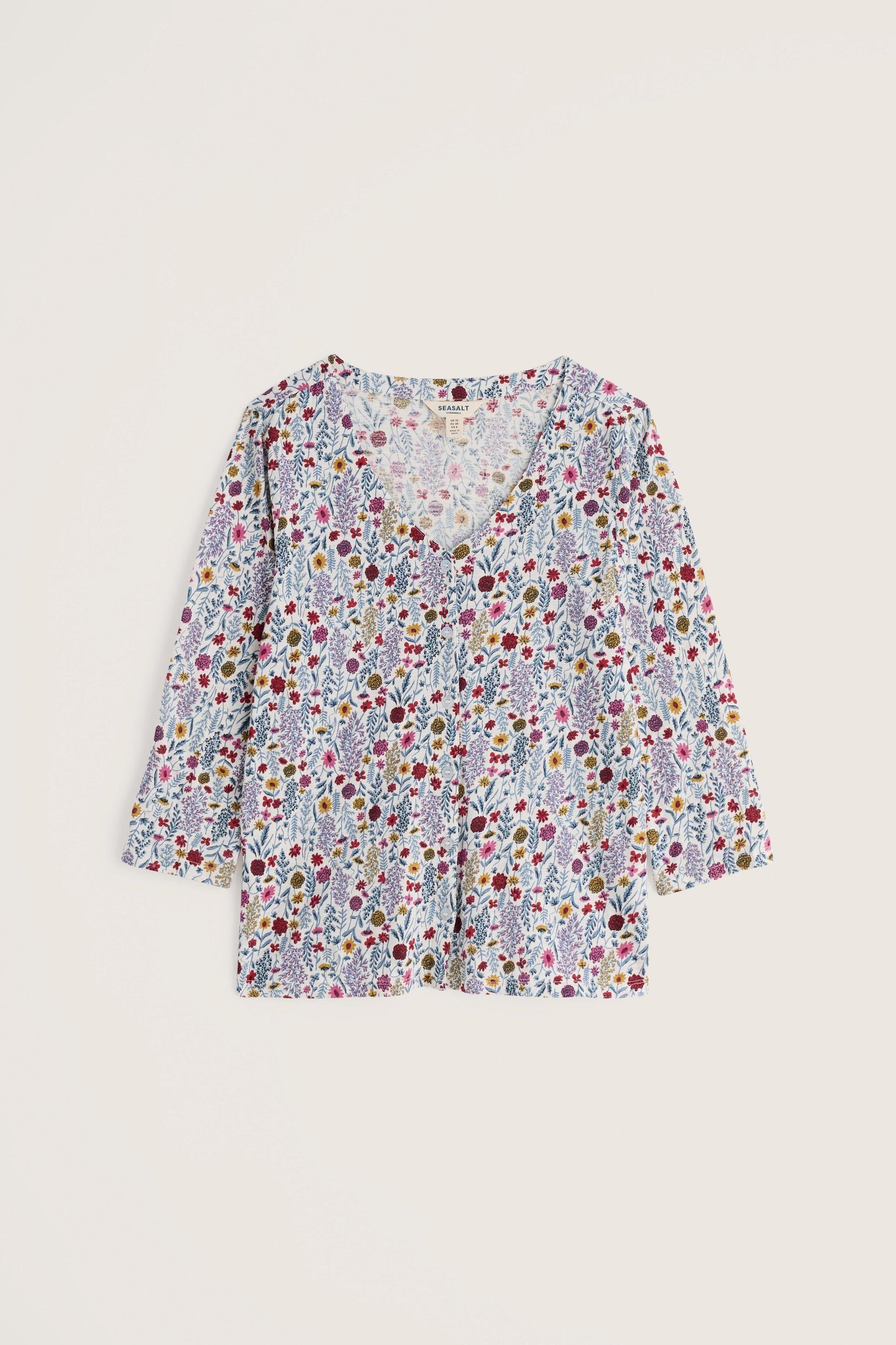 A long-sleeve V-neck blouse with a dense multicolor floral and botanical print on a light background, displayed flat against a solid light beige backdrop.