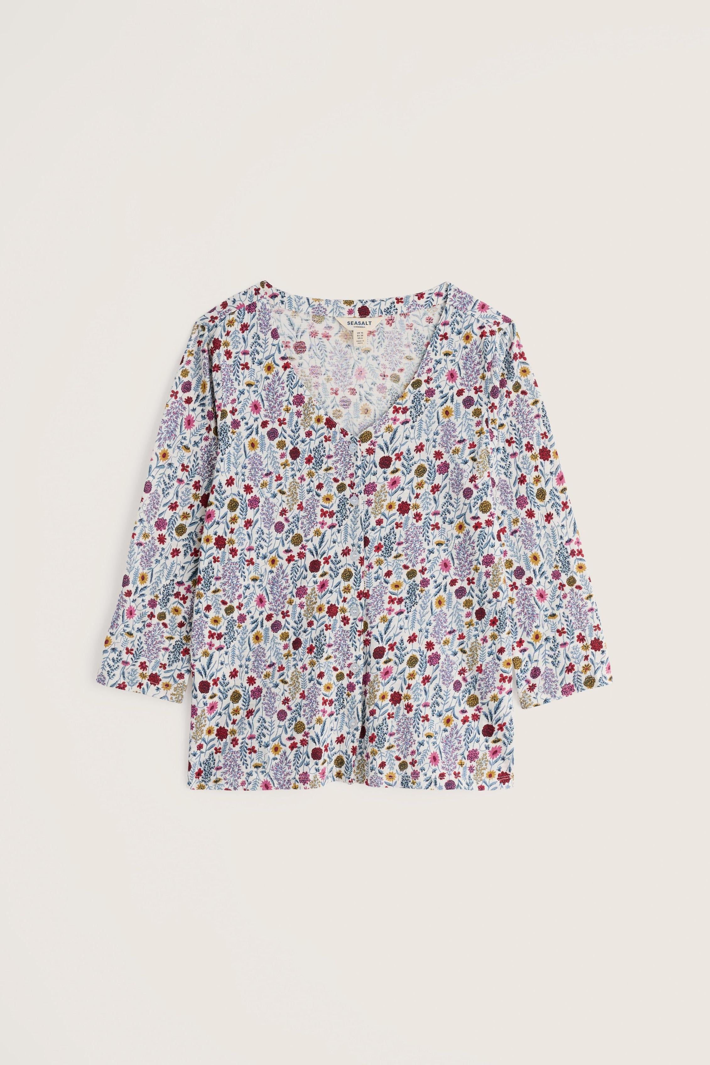 A long-sleeve V-neck blouse with a dense multicolor floral and botanical print on a light background, displayed flat against a solid light beige backdrop.