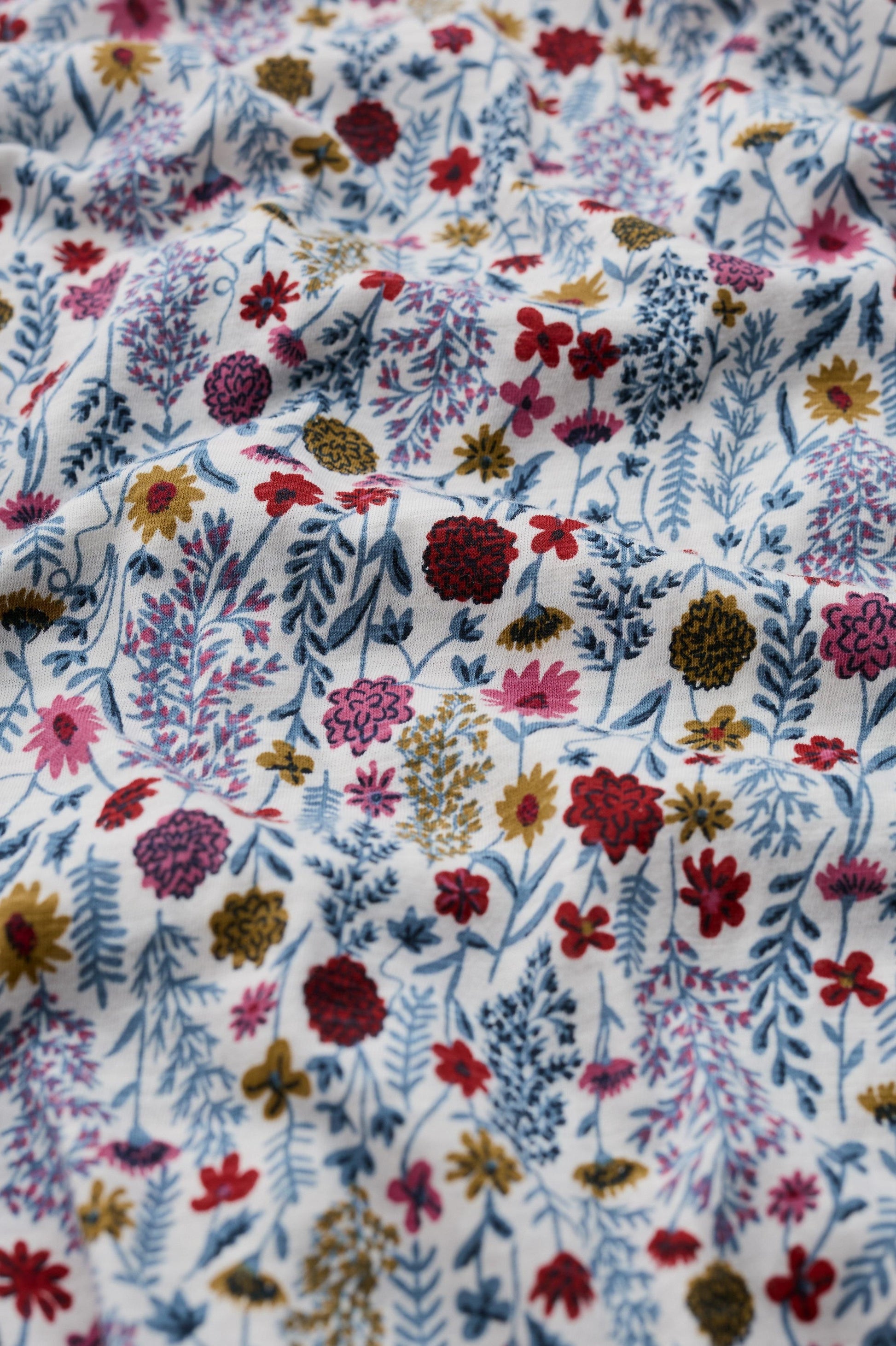 A close-up of a white fabric with a dense, multicolored floral and botanical print featuring red, pink, blue, purple, and yellow flowers and leaves.