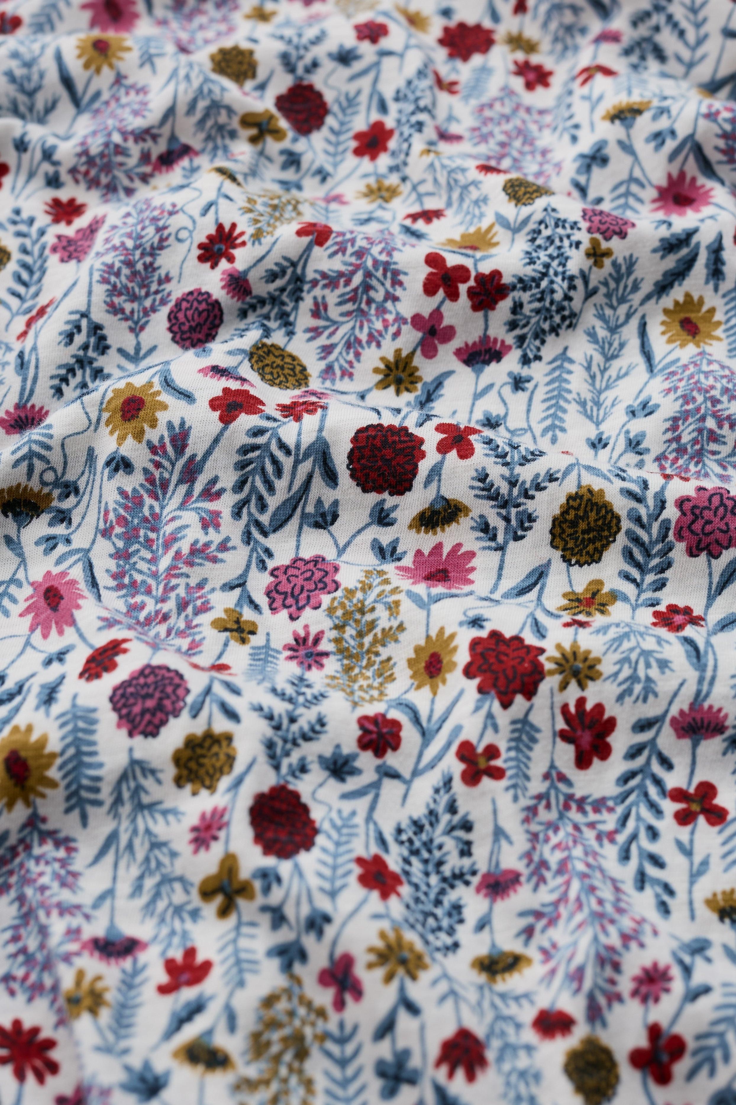 A close-up of a white fabric with a dense, multicolored floral and botanical print featuring red, pink, blue, purple, and yellow flowers and leaves.