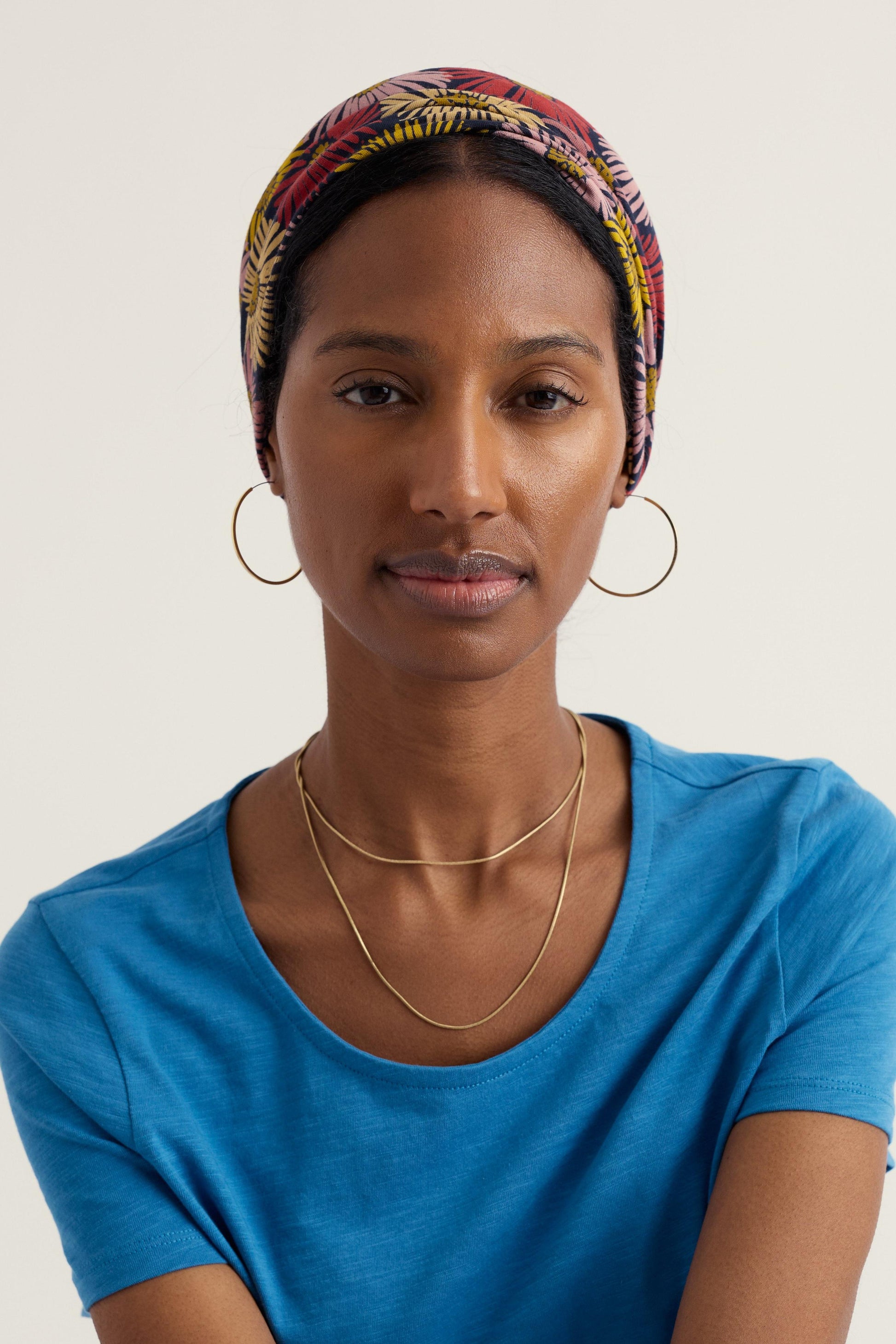 Woman wearing a colorful headscarf, blue shirt, and gold jewelry on a plain background