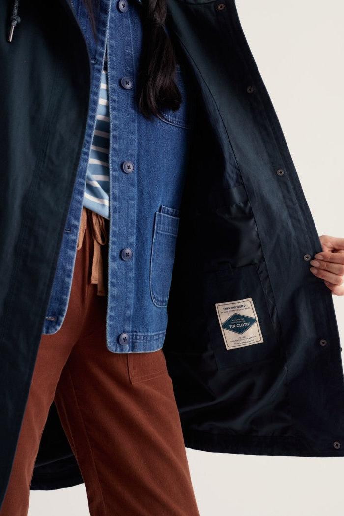 A close-up of a person wearing a dark navy coat over a blue denim shirt and rust-brown pants, holding the coat open to show the interior pocket with a sewn-in label.