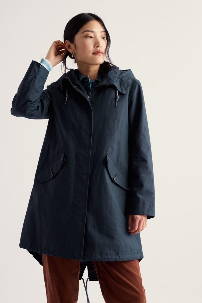 A woman wearing a dark navy hooded parka jacket with front flap pockets, styled over a teal shirt and rust-brown pants, photographed against a plain light beige studio background.