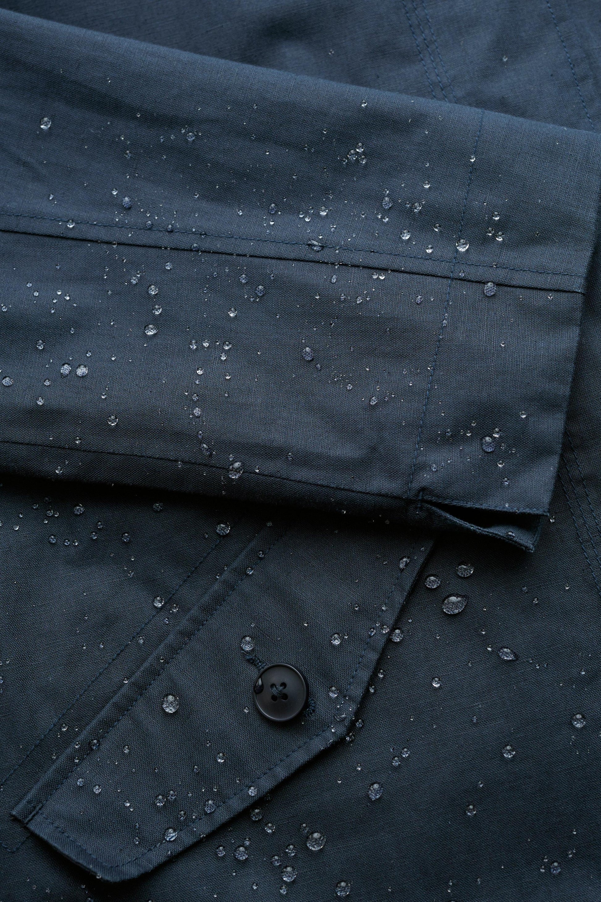 Close-up of a dark navy or charcoal fabric garment with water droplets beaded on the surface, showing a folded cuff and a buttoned pocket.