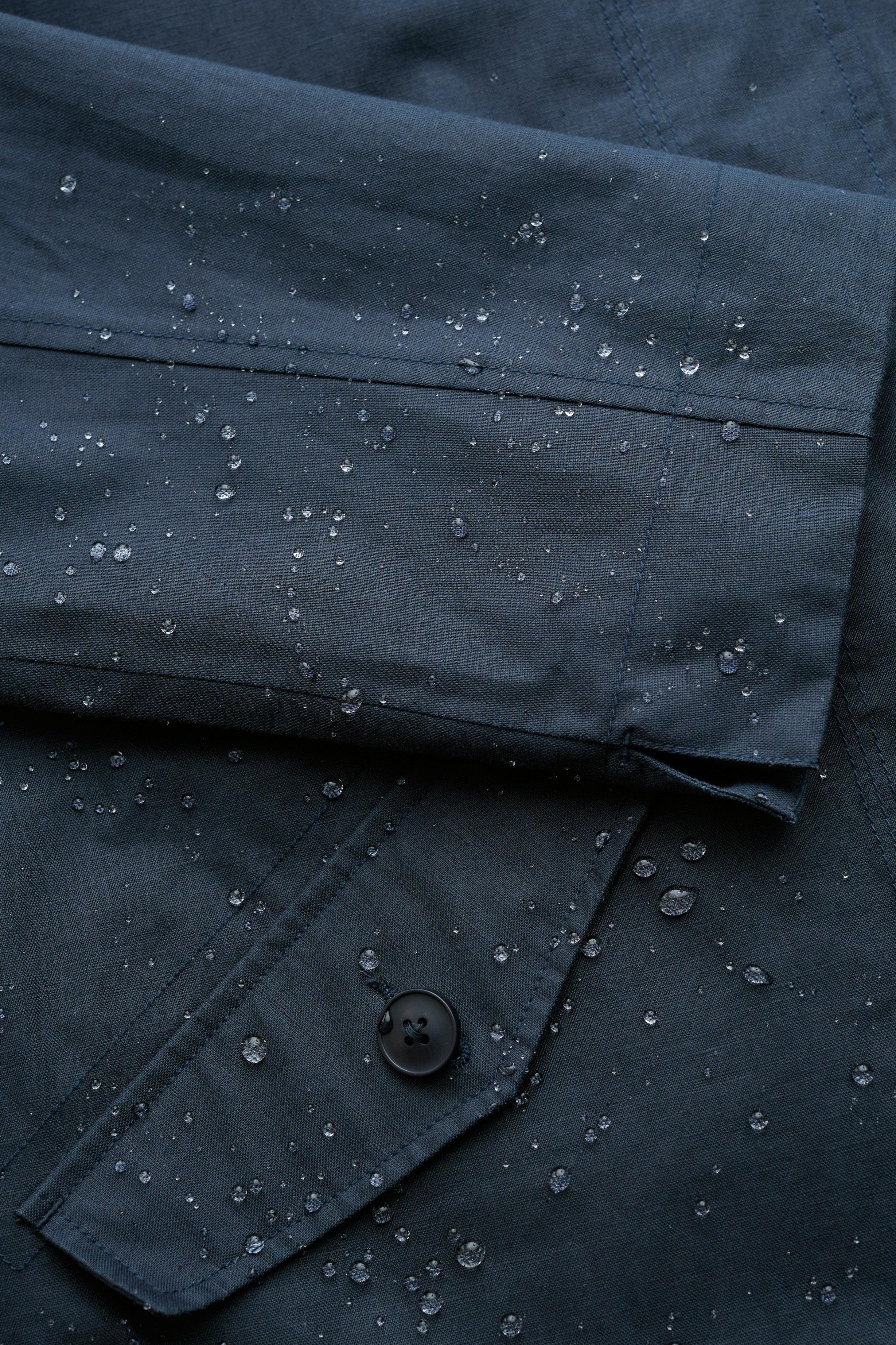 Close-up of a dark navy or charcoal fabric garment with water droplets beaded on the surface, showing a folded cuff and a buttoned pocket.