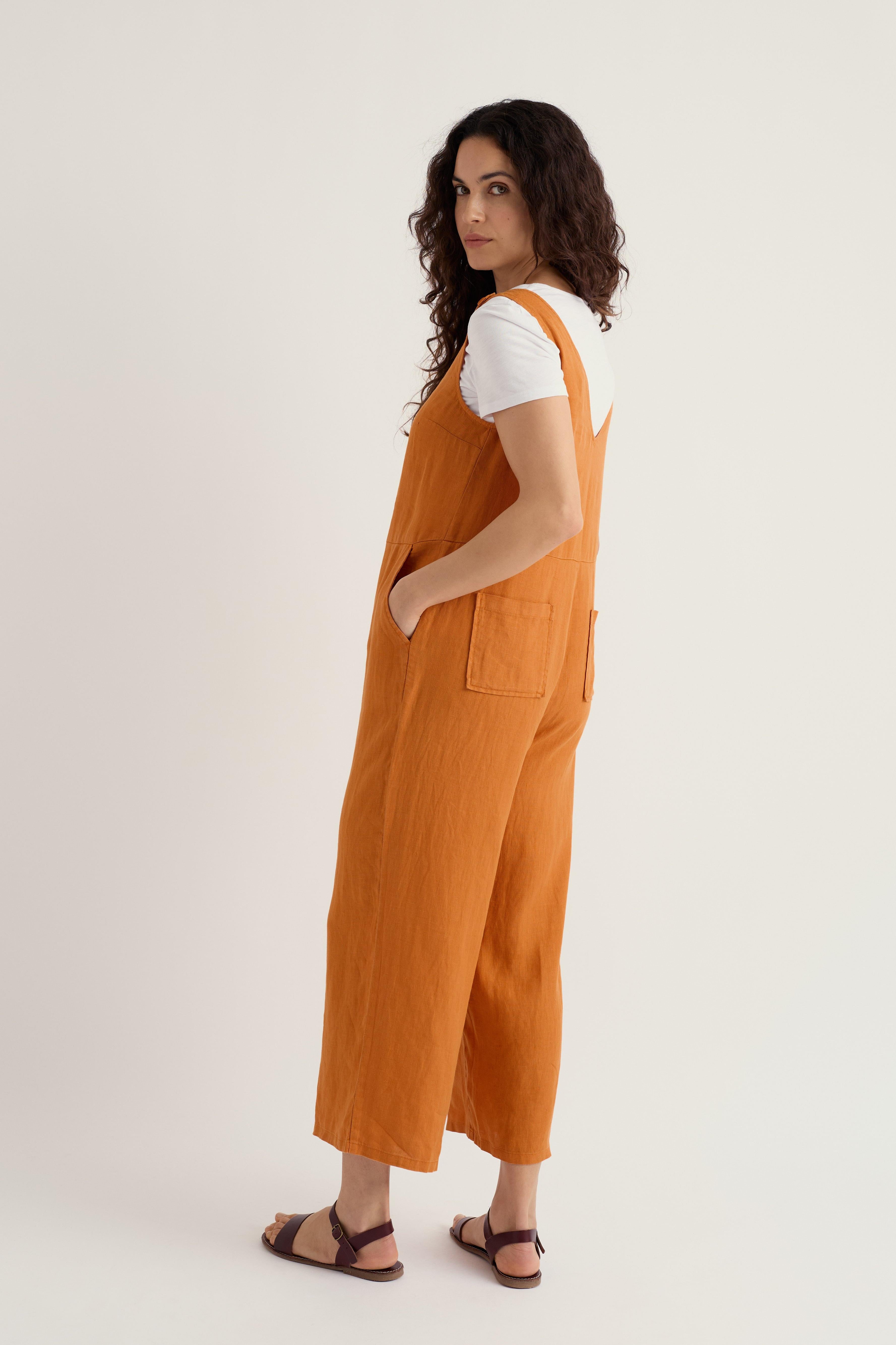 Woman standing in a casual orange linen jumpsuit and white t-shirt looking over her shoulder against a white background
