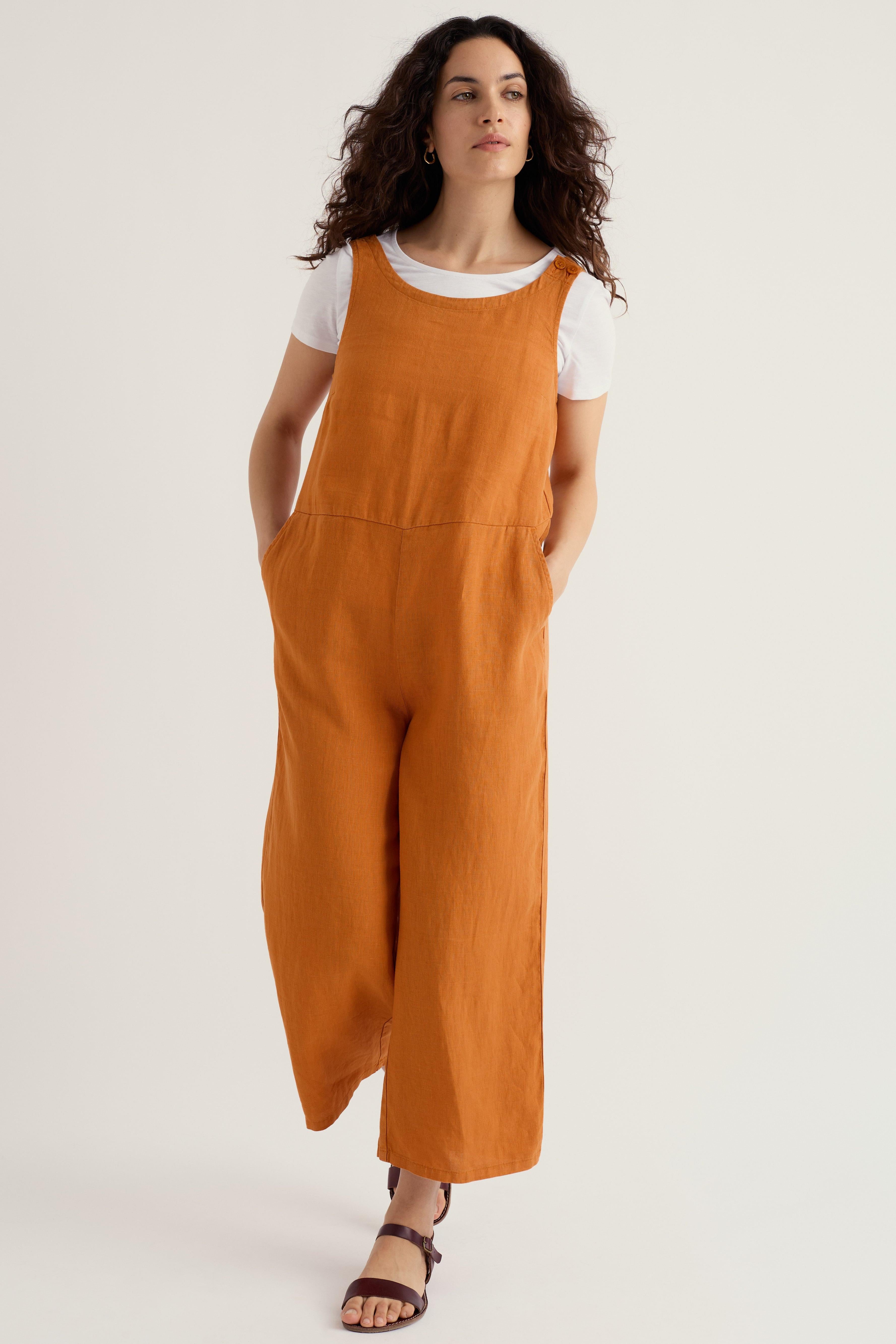 A woman with curly hair wearing a burnt orange linen jumpsuit over a white t-shirt against a light studio background.