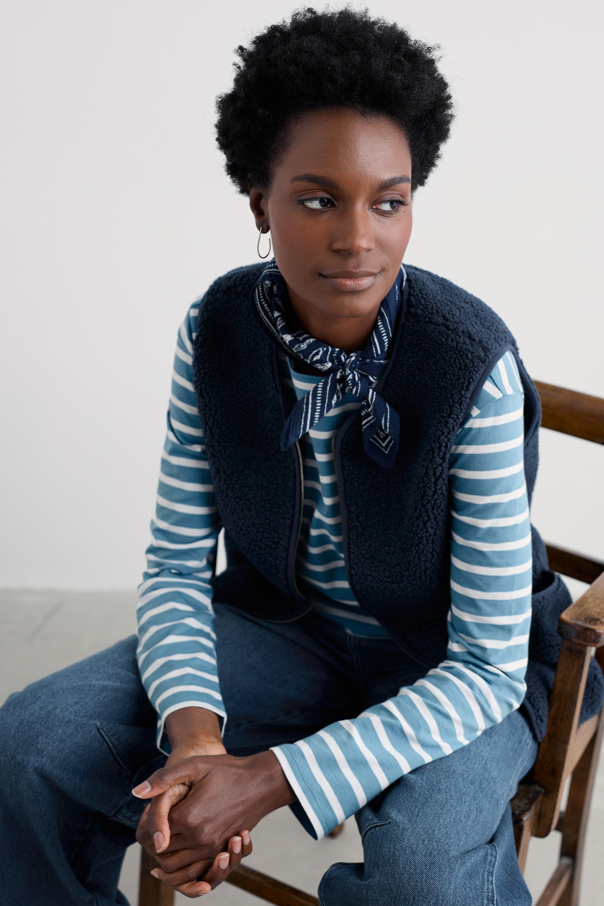 Woman wearing a blue and white striped shirt with a navy vest and jeans, sitting on a wooden chair.
