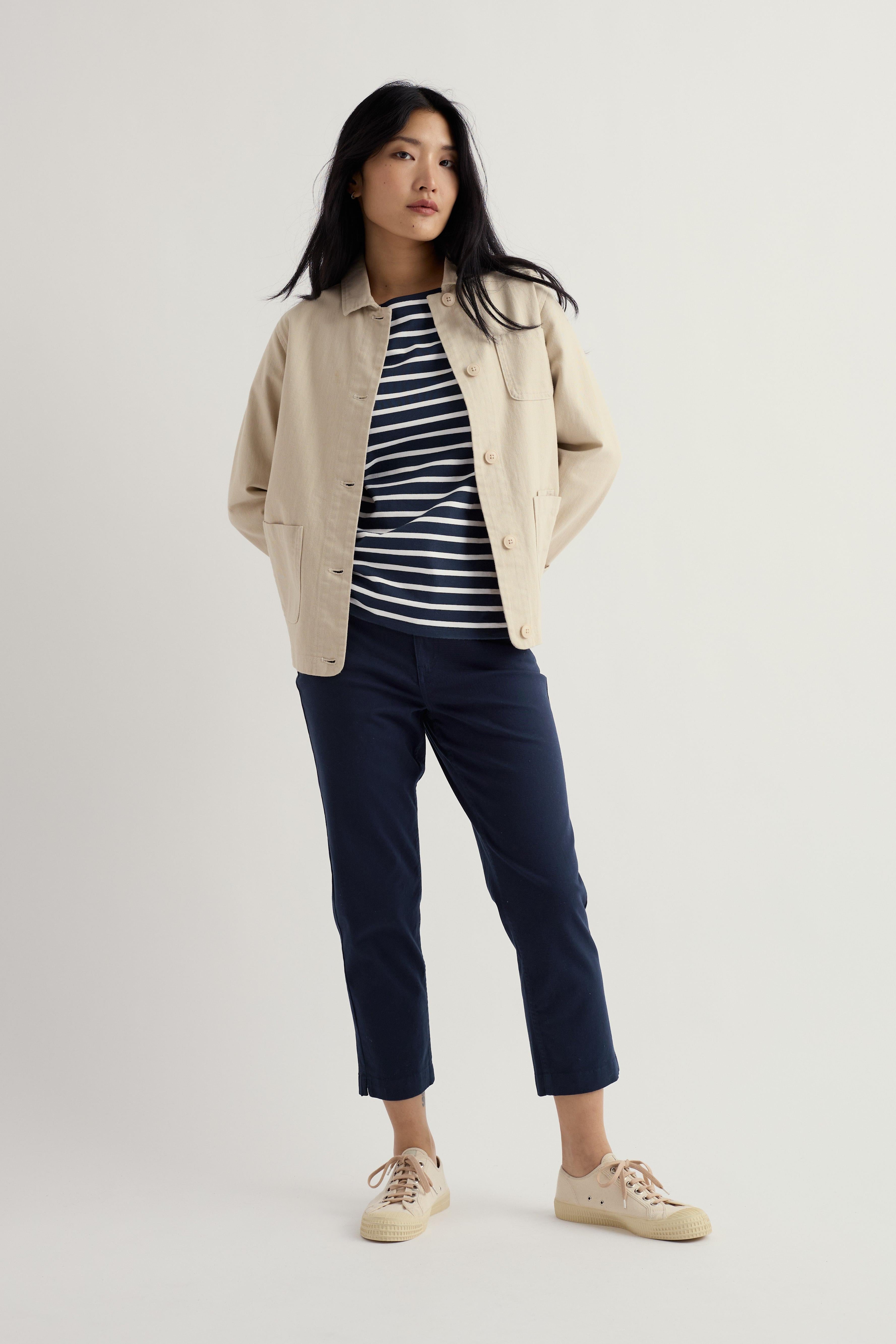 Woman wearing a beige jacket, striped shirt, navy organic cotton trousers, and beige shoes on a white background