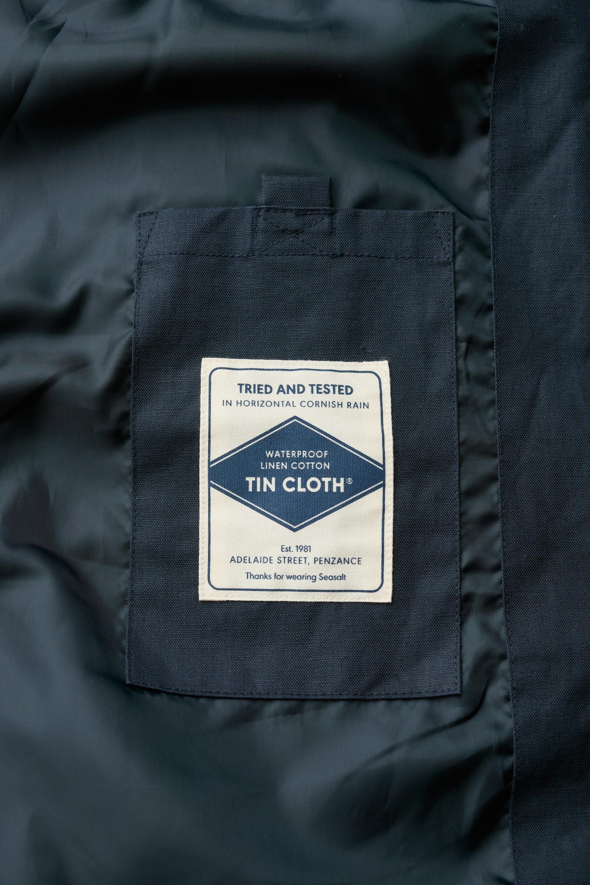 Close-up of a dark navy blue coat showing a sewn-in label patch on the chest pocket. The white rectangular label reads 'Tried and Tested in Horizontal Cornish Rain' and 'Waterproof Linen Cotton Tin Cloth'.
