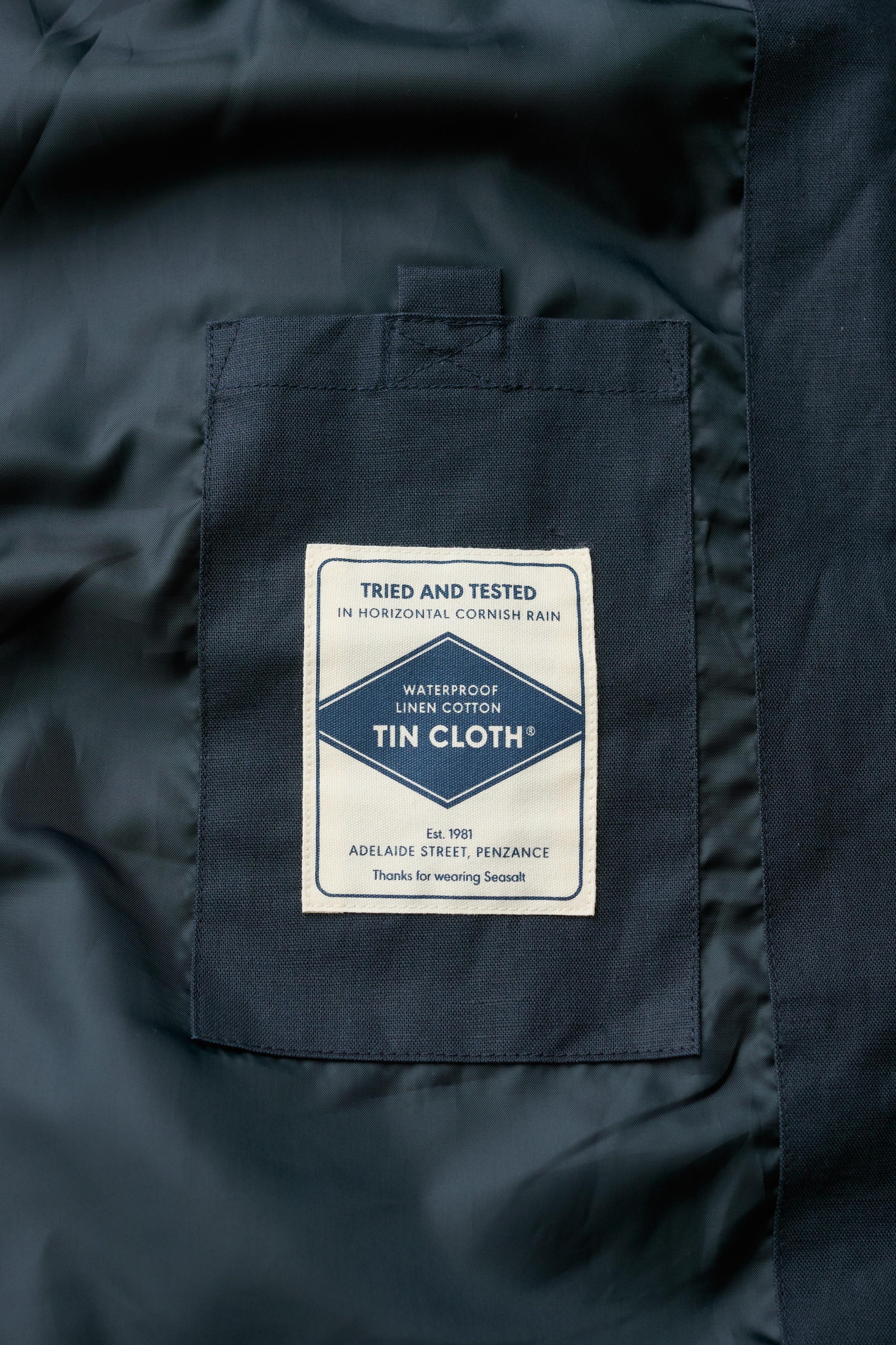 Close-up of a dark navy blue coat showing a sewn-in label patch on the chest pocket. The white rectangular label reads 'Tried and Tested in Horizontal Cornish Rain' and 'Waterproof Linen Cotton Tin Cloth'.