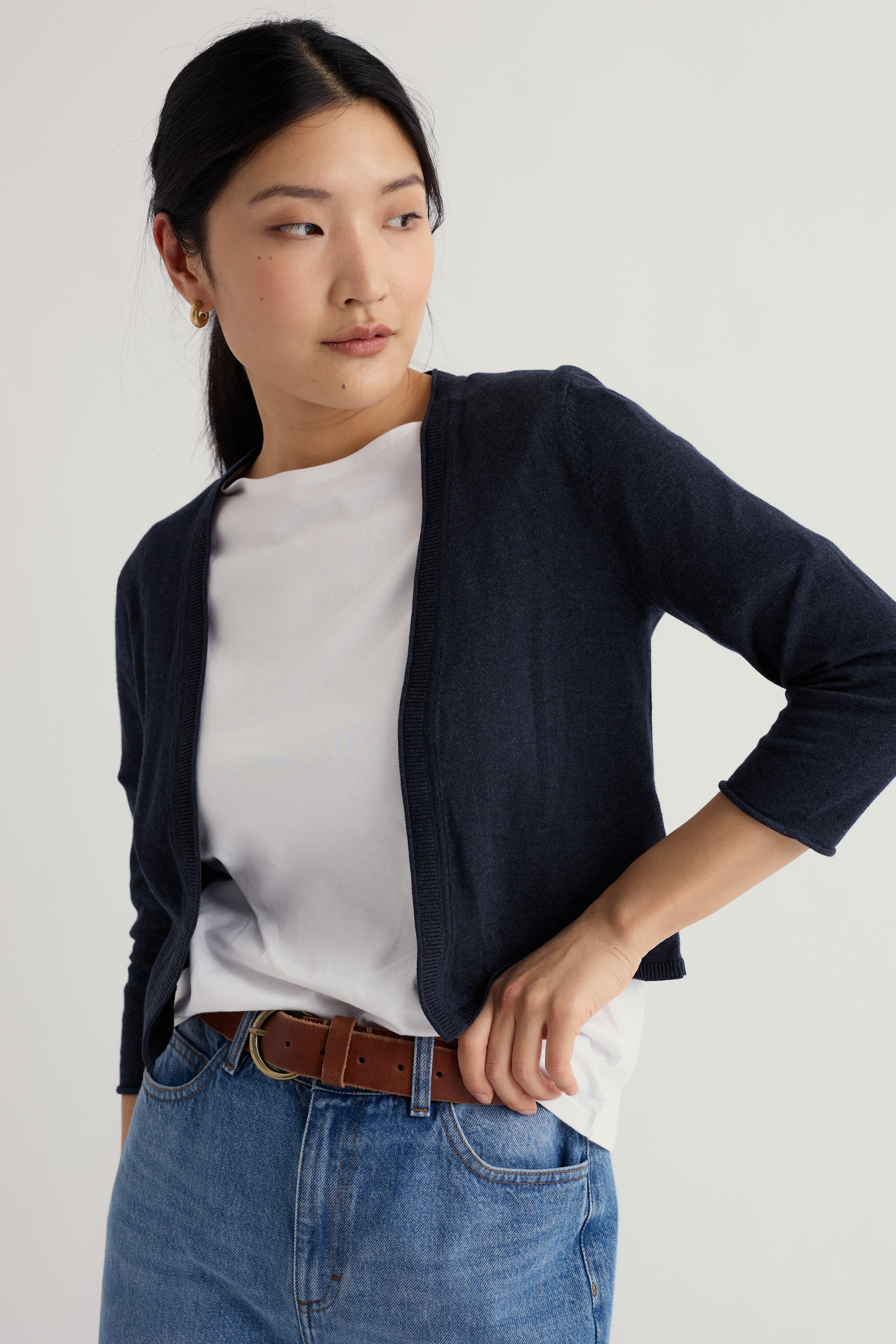 Woman wearing a navy organic cotton cardigan, white shirt, and blue jeans on a light gray background