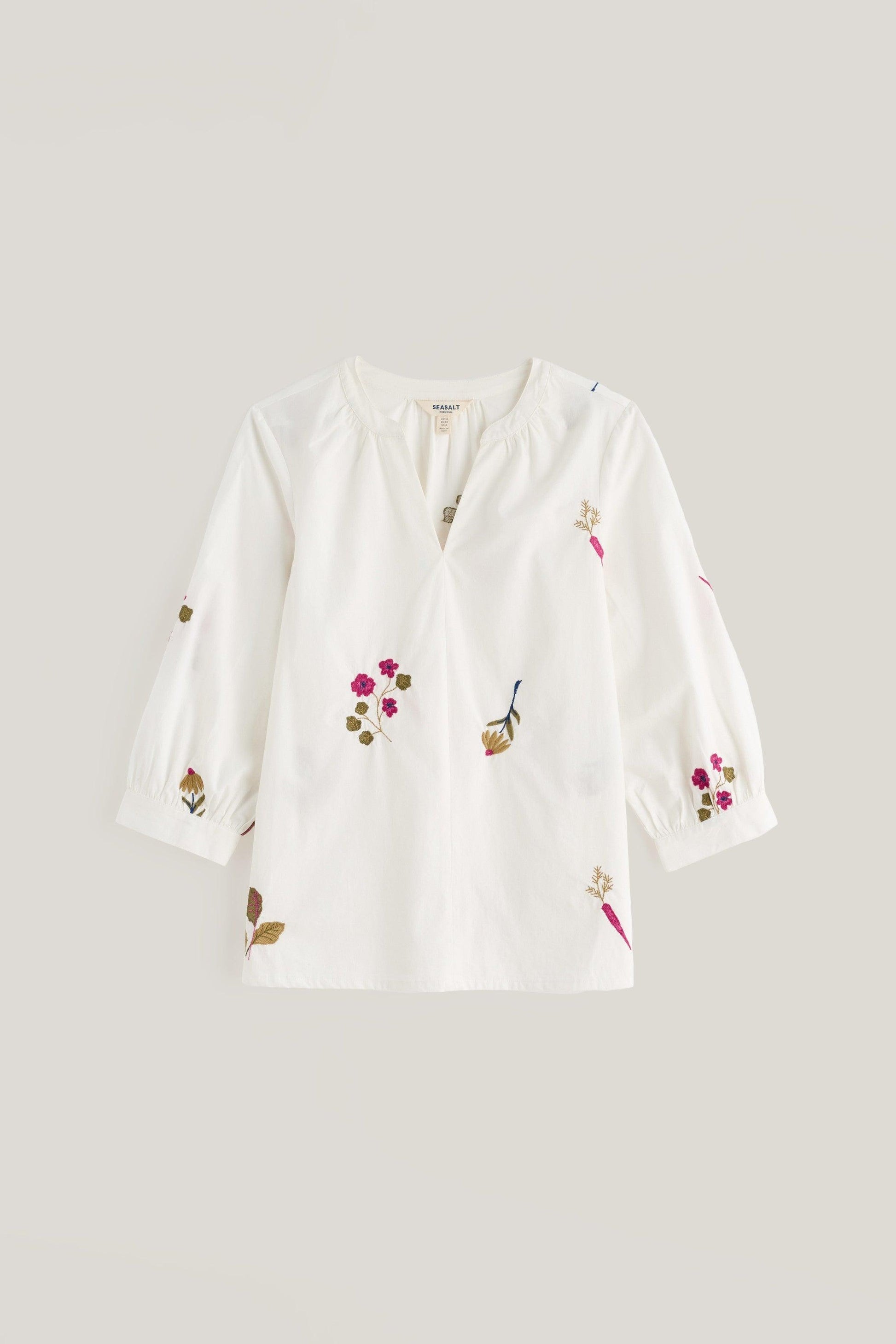 An off-white long-sleeve blouse with delicate floral and botanical embroidery in pink, green, and brown, displayed against a solid light gray background.