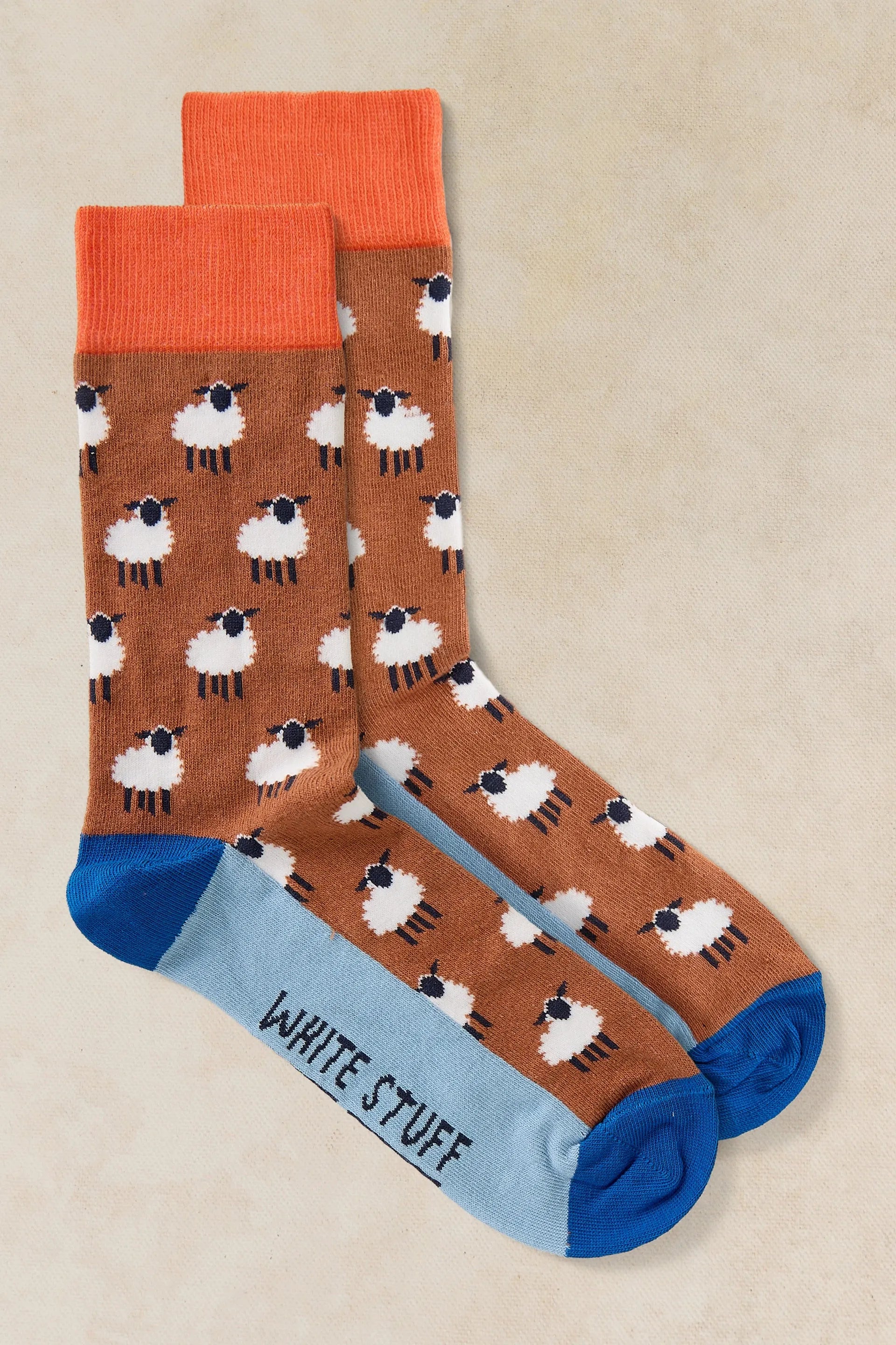 A pair of crew socks with a brown background patterned with white sheep, bright orange cuffs, light blue heels, and blue toes. The word 'WHITE STUFF' is printed in black on the light blue heel area.