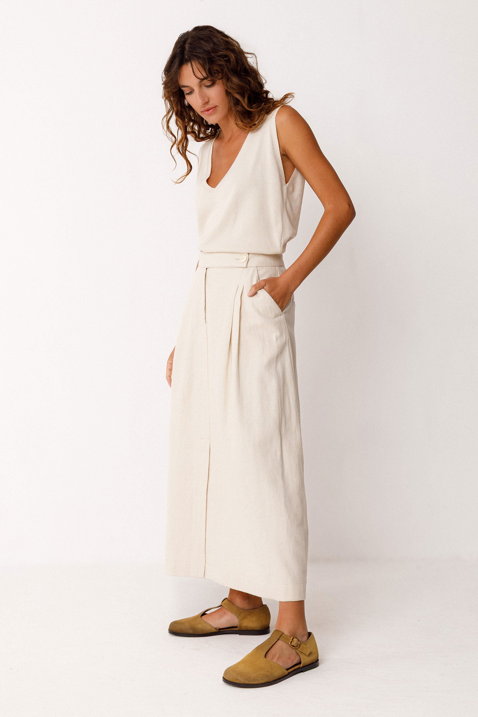 Woman wearing an ivory sleeveless top and an a-line ivory midi skirt with brown shoes on a white background