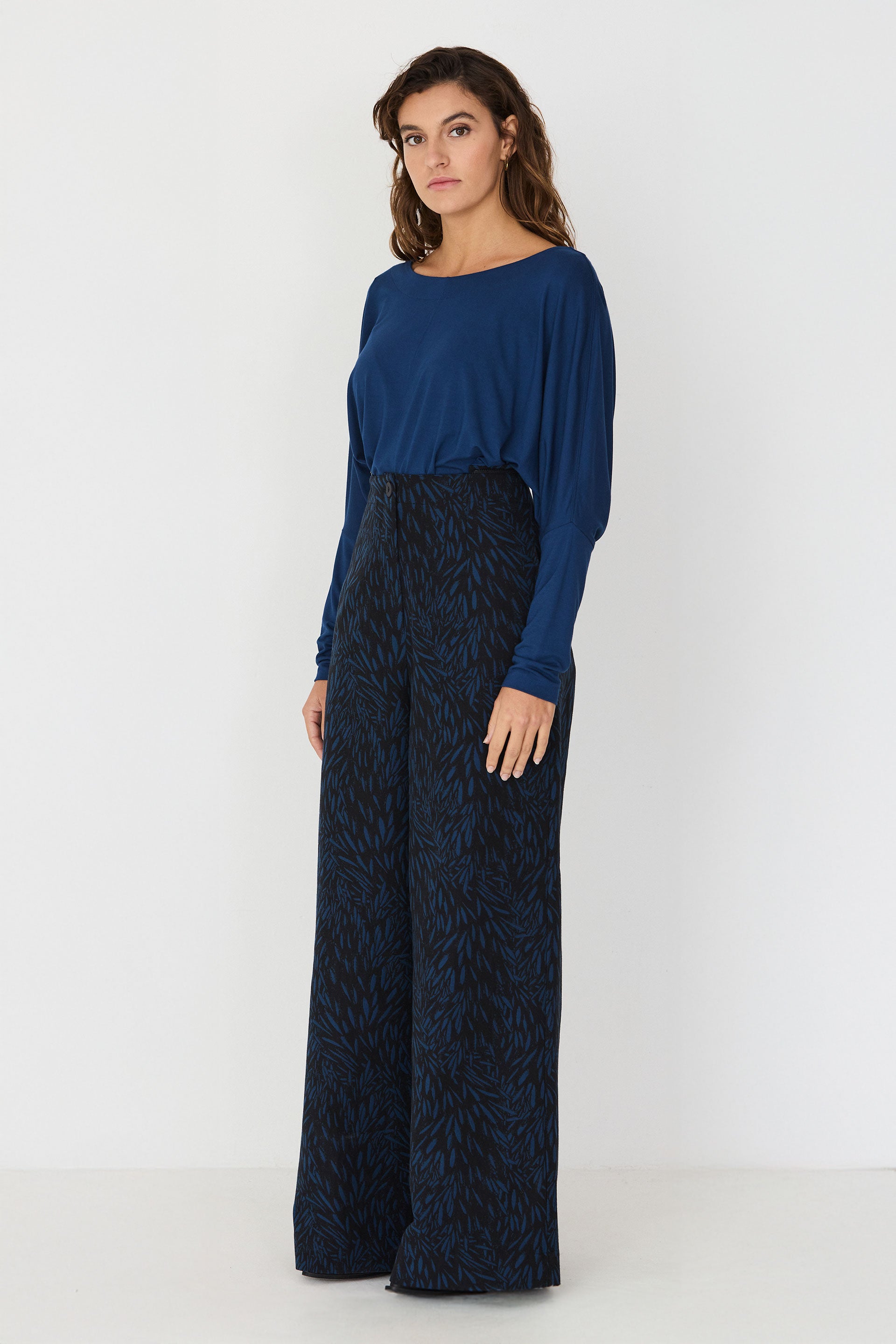 A model standing wearing a blue long-sleeve top and patterned wide-leg trousers that form a two-piece blue jumpsuit.