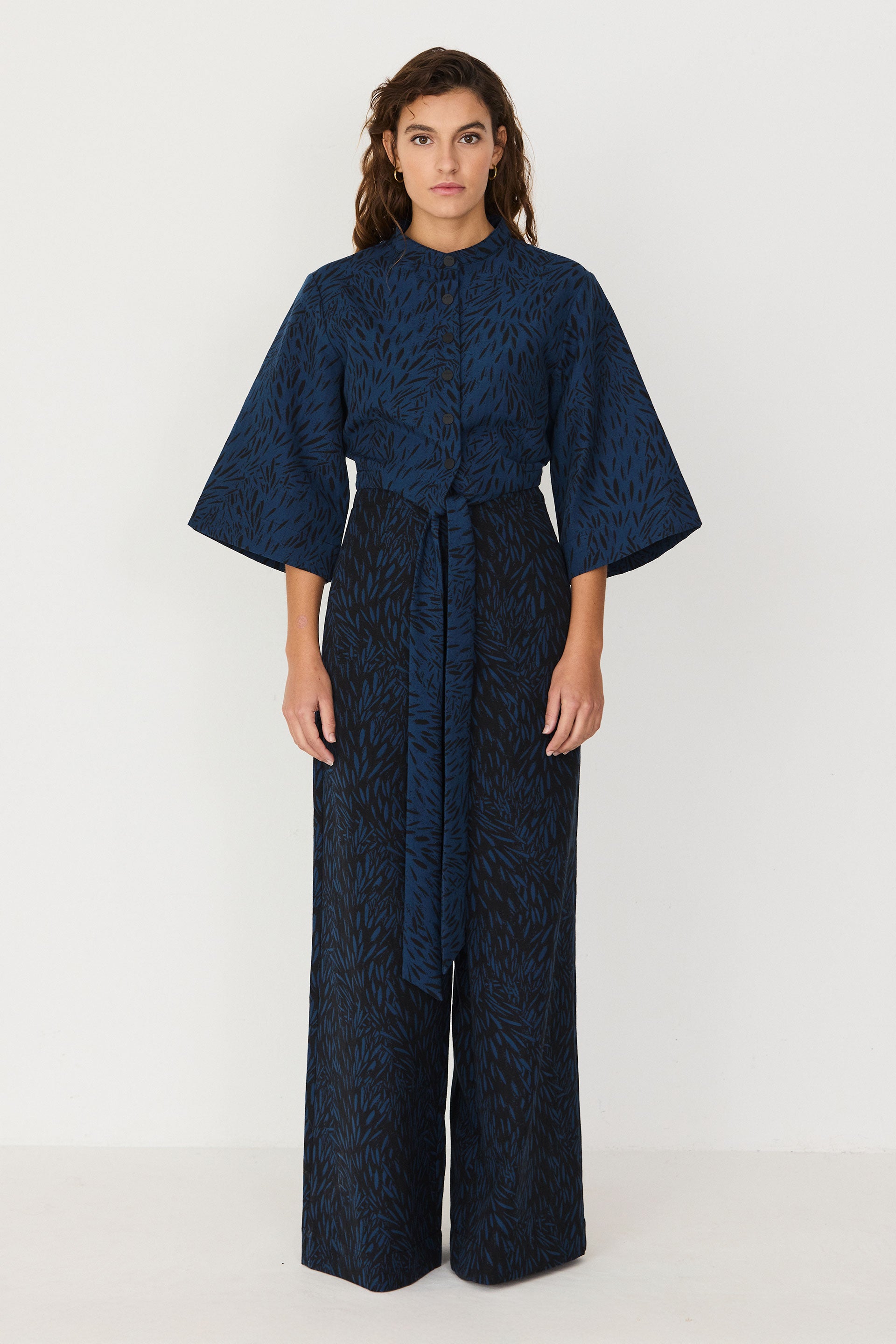 A woman wearing a blue organic cotton sustainable two-piece style jumpsuit with wide sleeves and a tie waist.