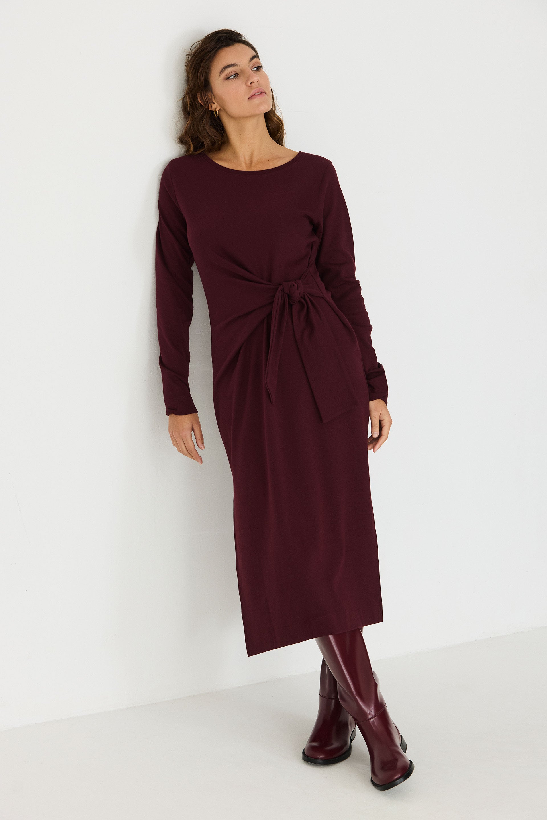 Woman leaning against a wall wearing a long-sleeved chocolate brown organic cotton midi dress by SKFK.