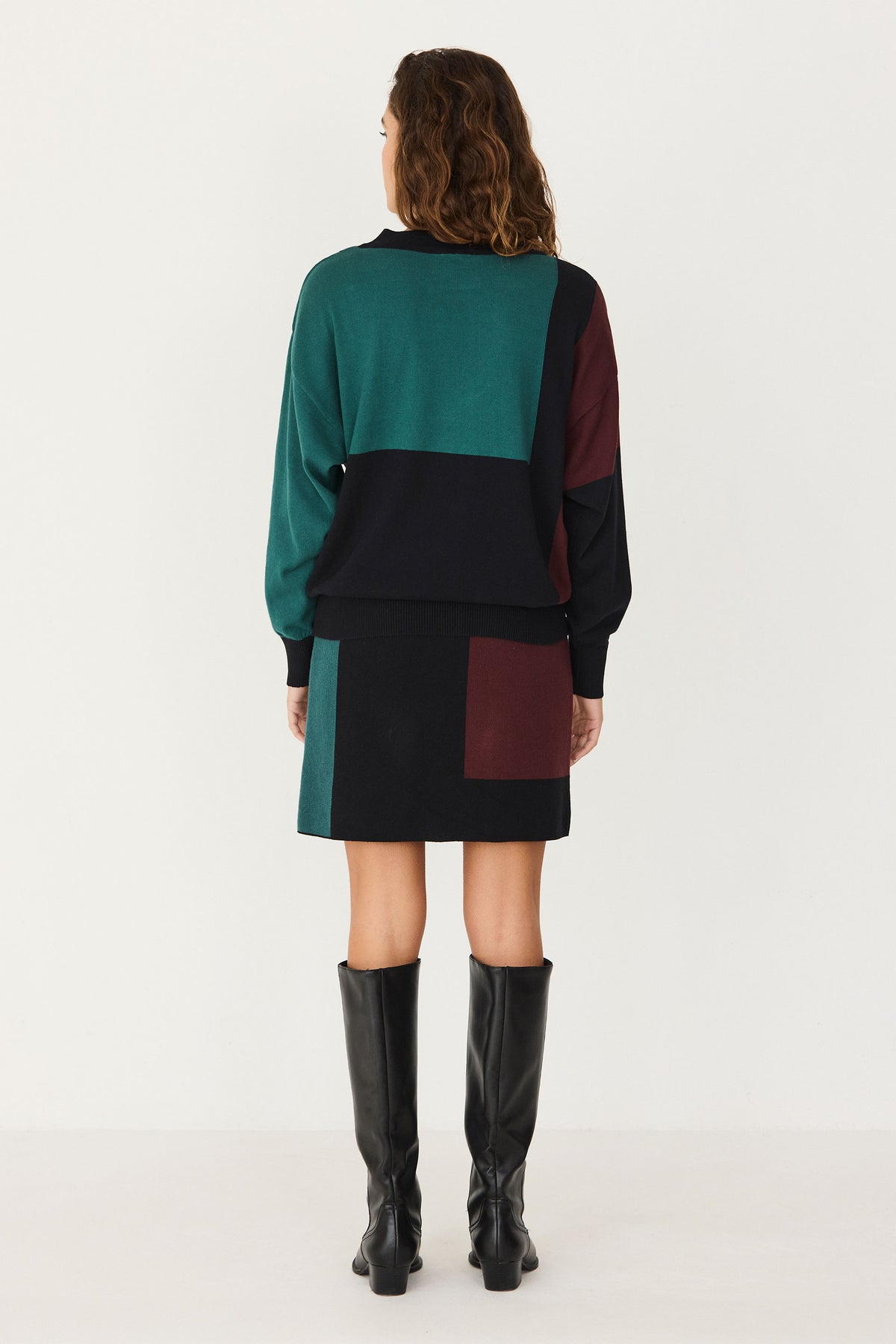 A woman wears a colourful cotton knitwear coord set consisting of a jumper and skirt, paired with black knee-high boots.