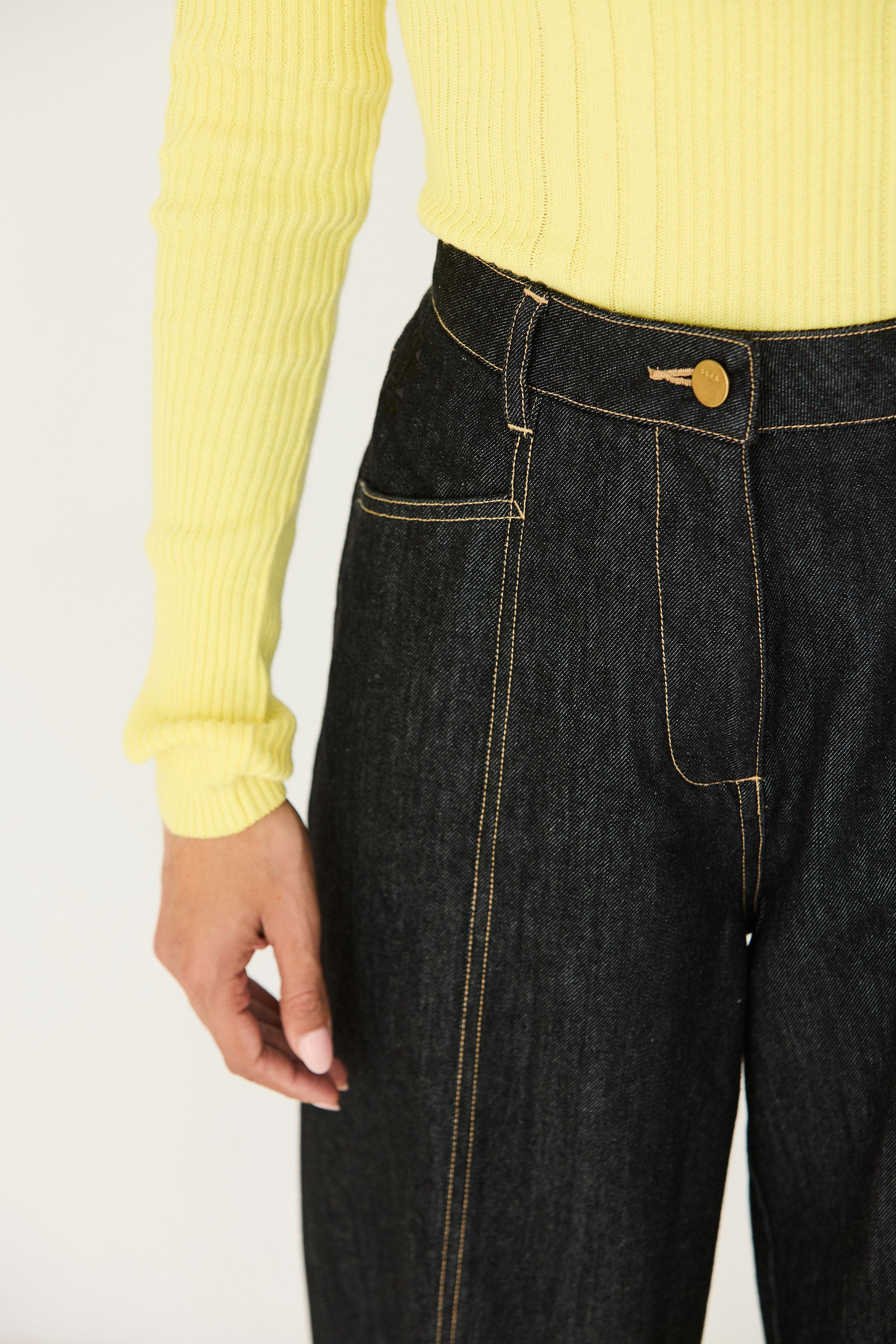A close-up view of a model wearing high-waisted black barrel leg jeans with contrasting stitching and a yellow sweater.