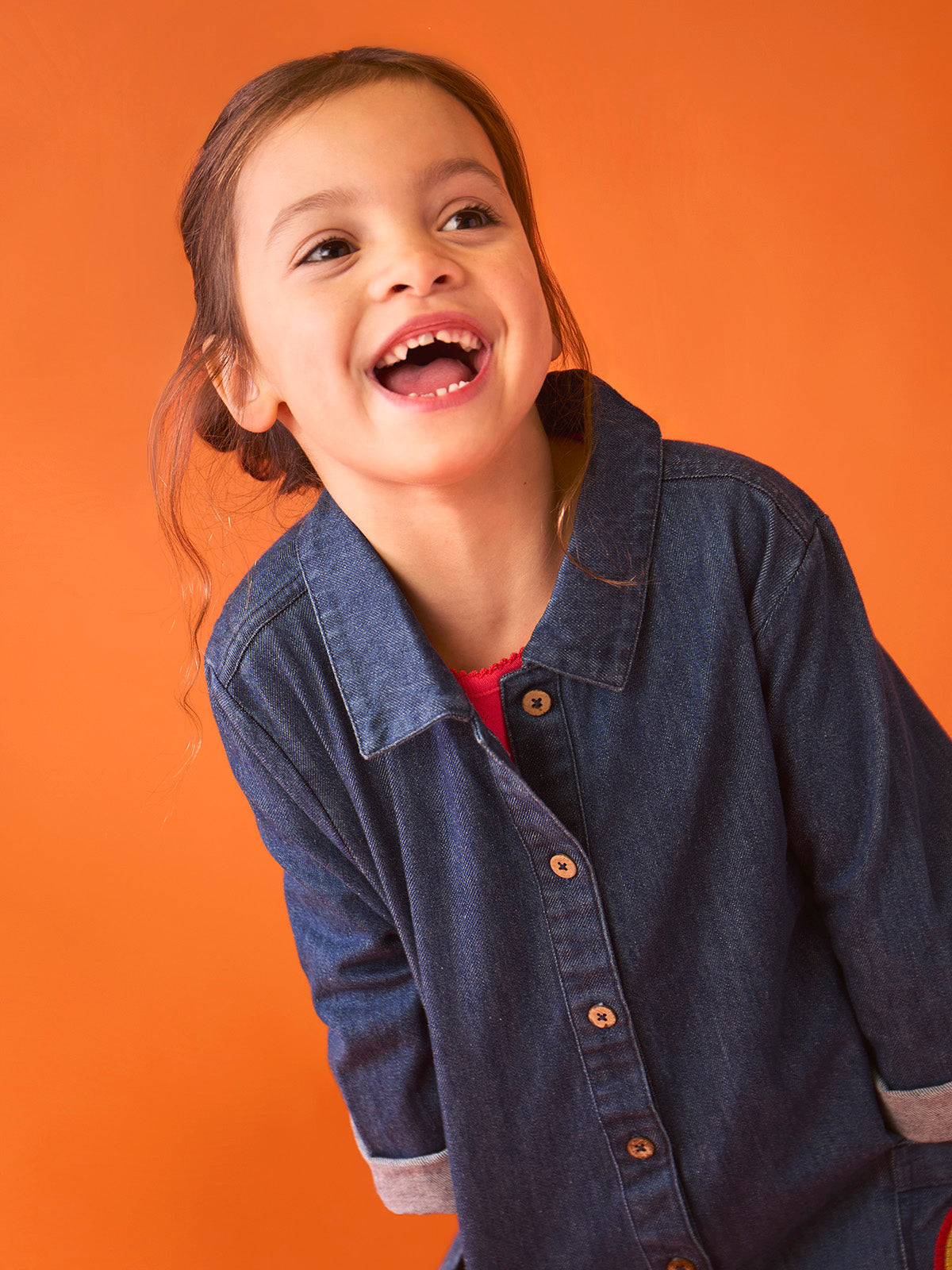 A happy young girl laughing while wearing a dark blue organic cotton denim jumpsuit against an orange background.