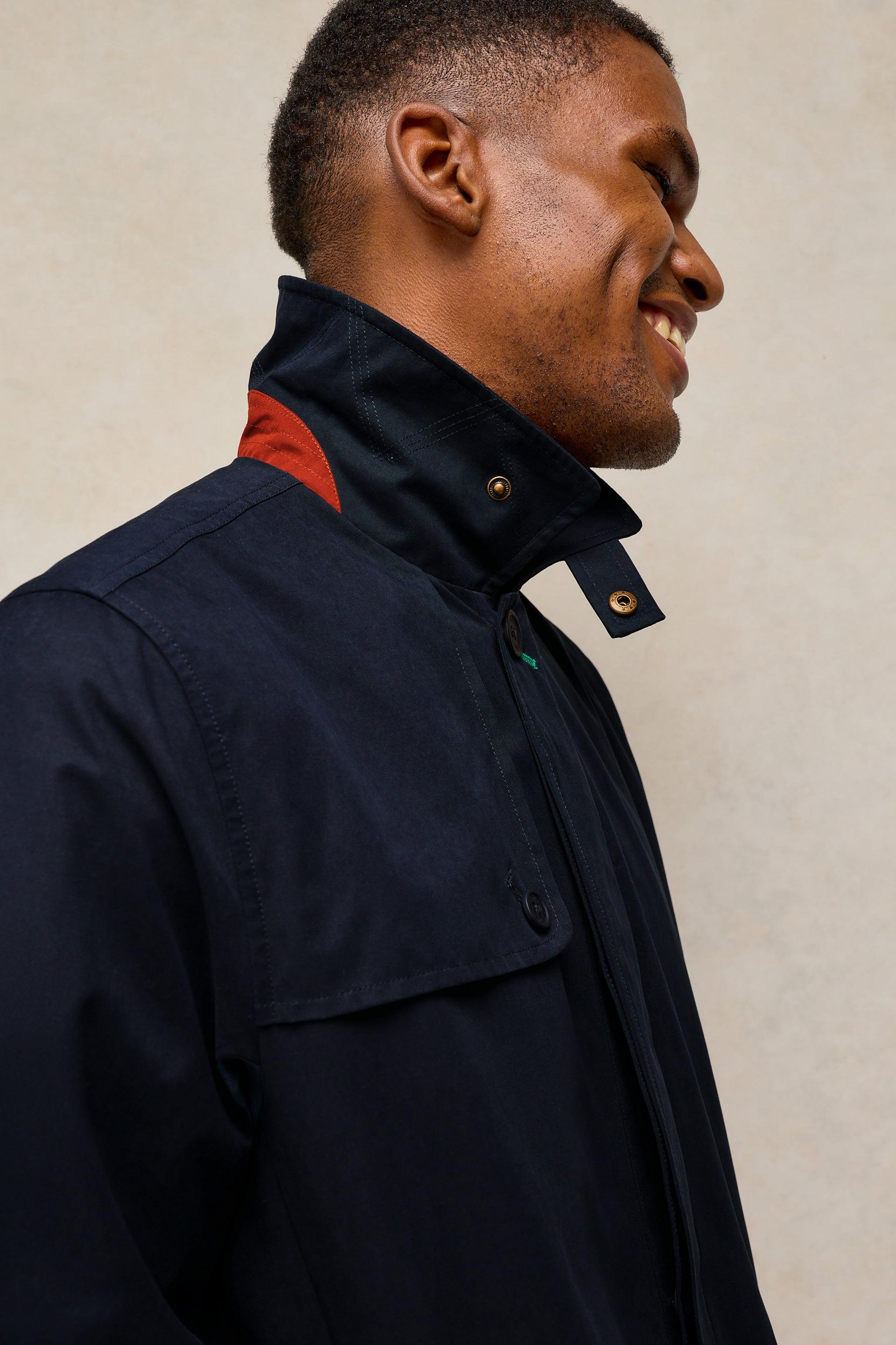 A close-up side profile of a man wearing a dark navy blue jacket with a high collar and a small red accent visible at the inner neck, set against a neutral beige background.