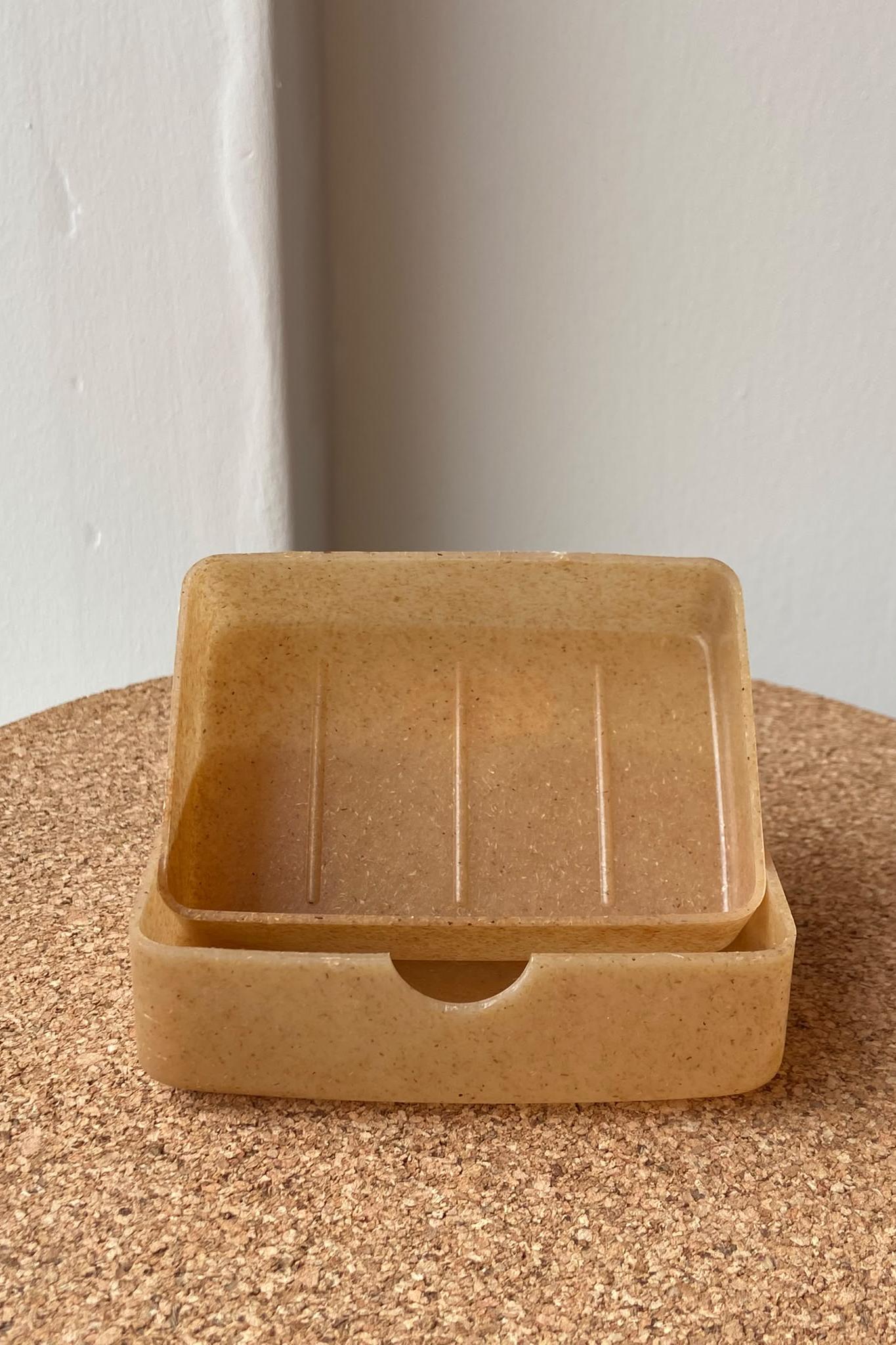A small square soap tray with a shallow front slot, shown nested on top of a matching base on a cork-like surface against a plain light wall.