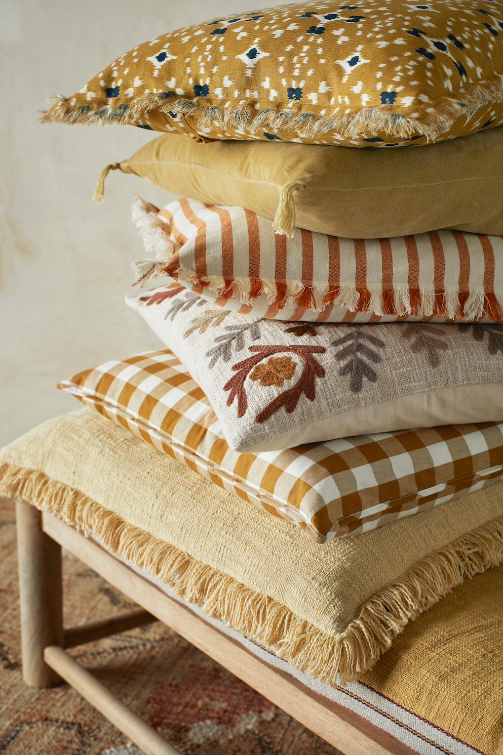 A tall stack of decorative cushions with patterns, fringes, and tassels on a wooden bench.