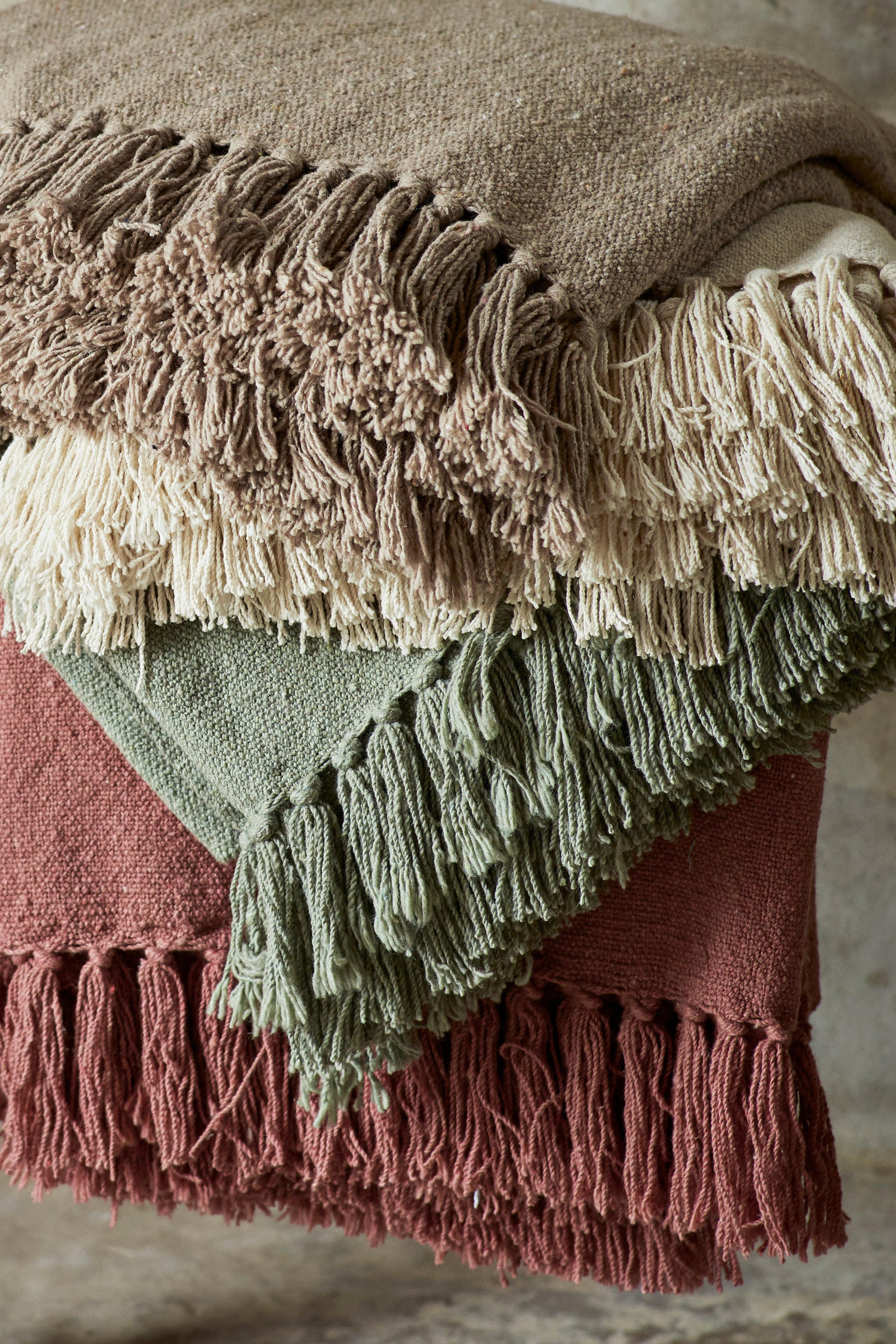 A stack of folded textured throw blankets with fringe edges in earthy tones of brown, cream, sage green, and terracotta.