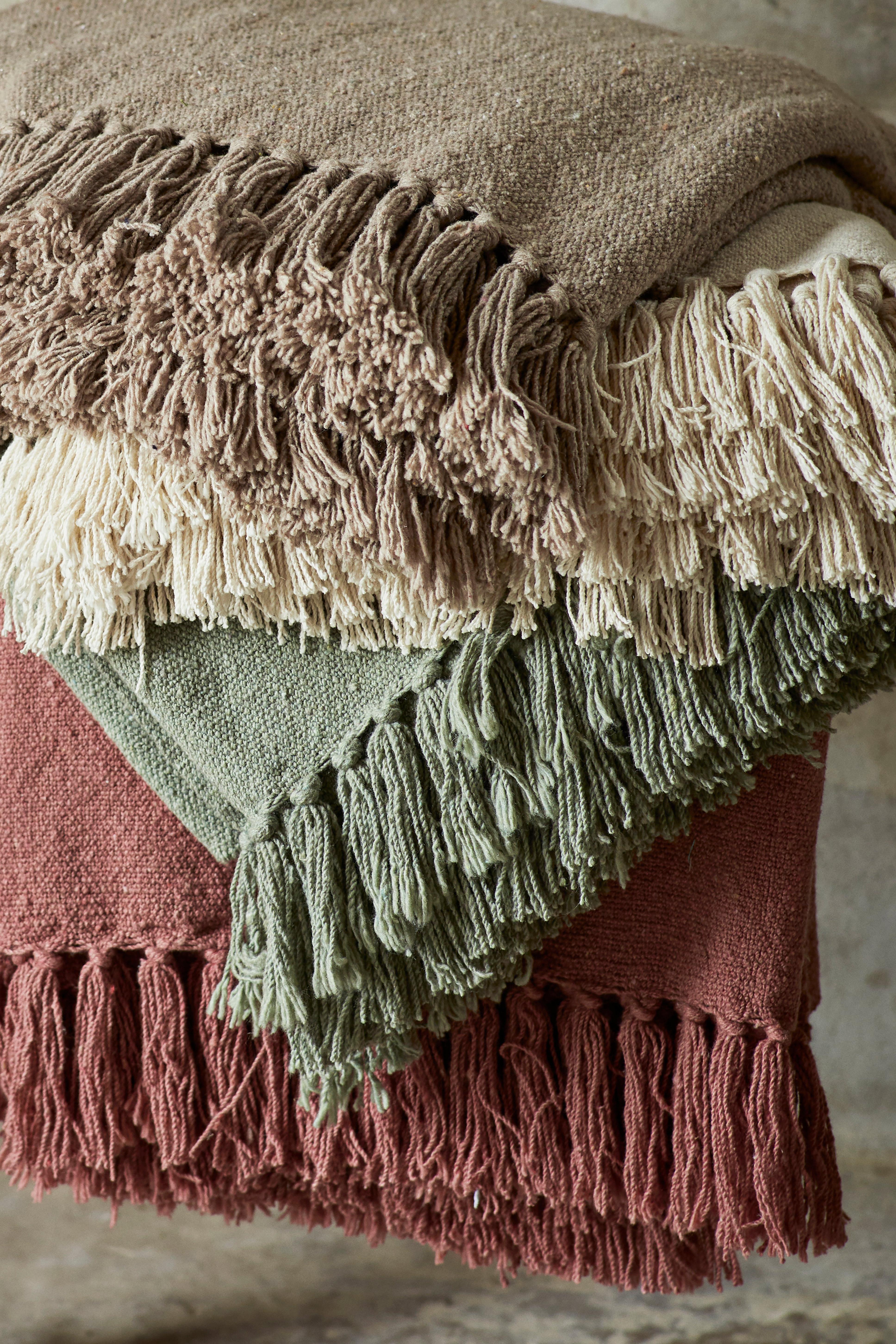 A stack of folded textured throw blankets with fringe edges in earthy tones of brown, cream, sage green, and terracotta.