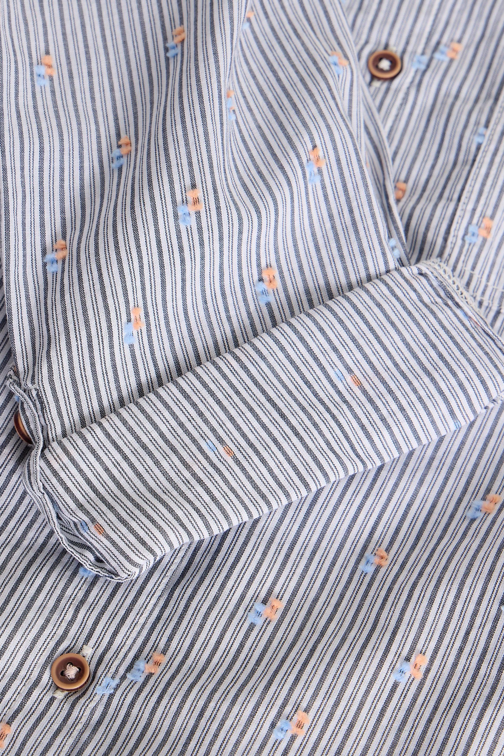 Close-up of a striped shirt fabric with vertical blue, gray, and white stripes, accented with small orange and light blue embroidered motifs and brown buttons.