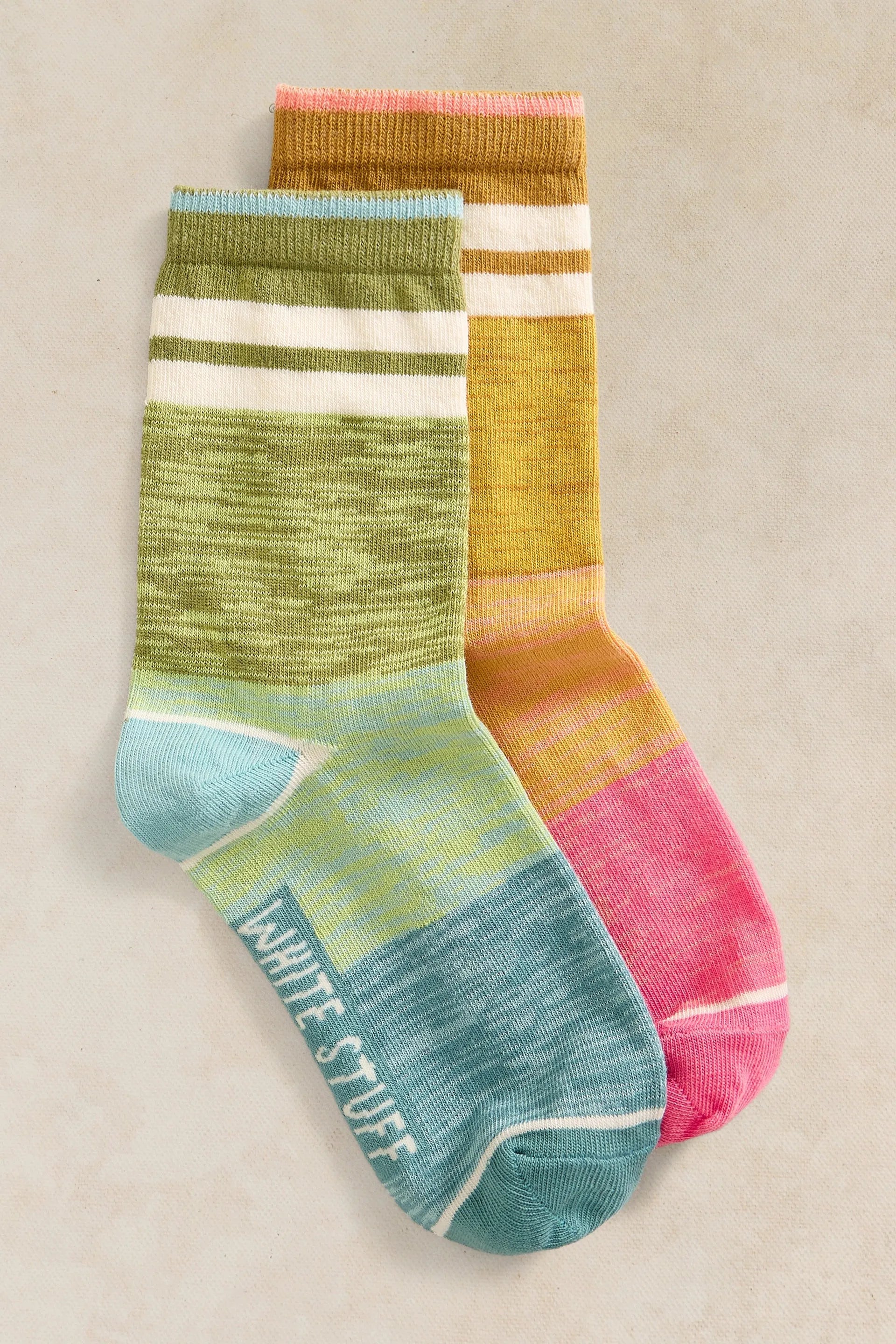 Two crew socks with horizontal color-blocked stripes in green, white, yellow, orange, and pink, with the brand name 'WHITE STUFF' printed on the toe of the front sock, displayed on a neutral beige background.