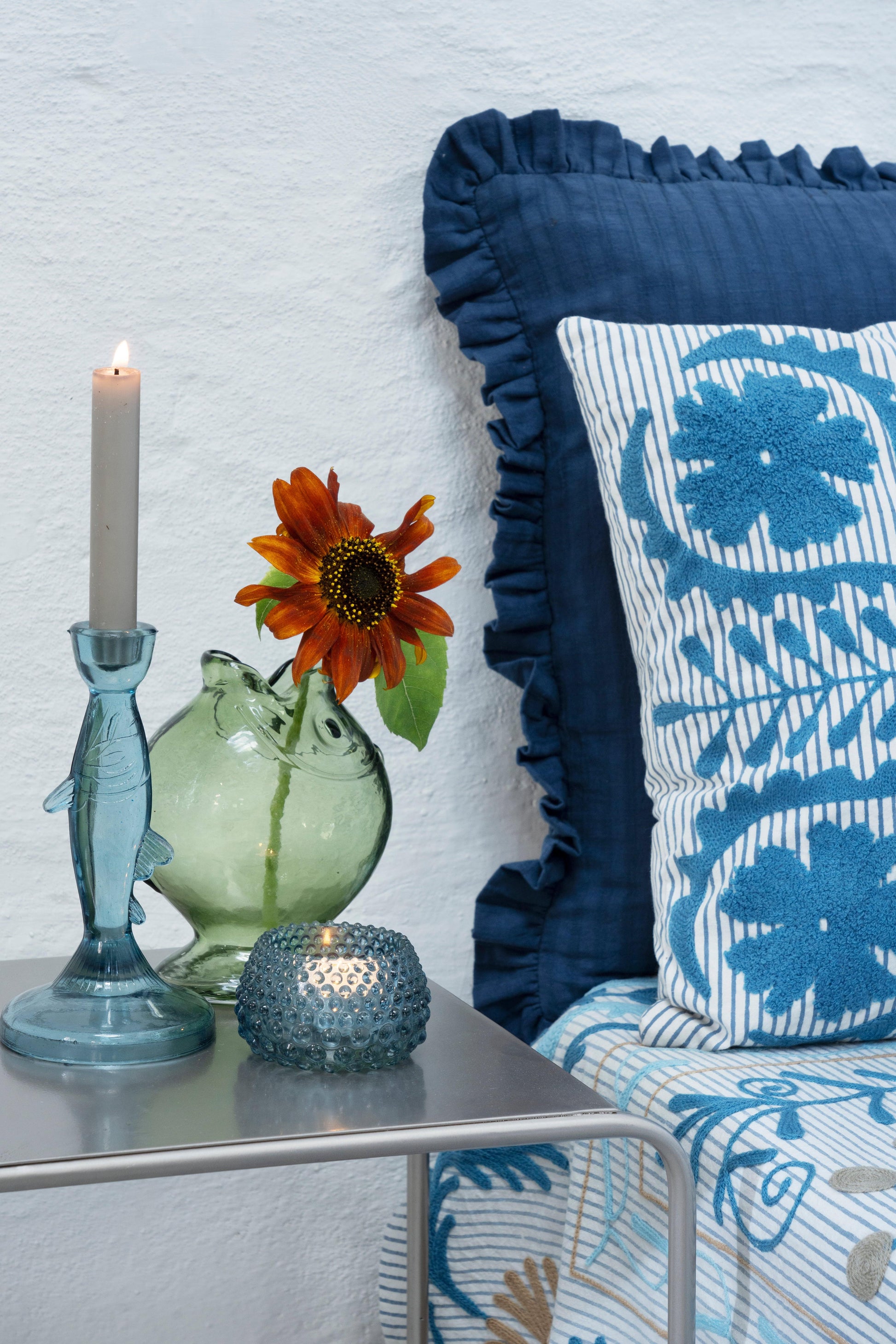 A vibrant sunflower in a green glass vase beside a lit candle on a bedside table with blue patterned pillows.