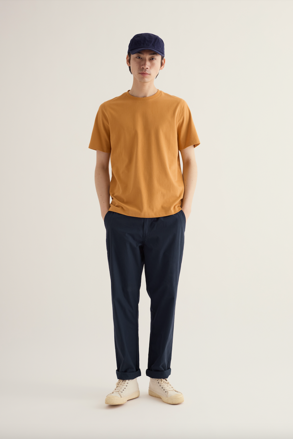 A man wearing a mustard organic cotton mens t-shirt, dark trousers, and a navy baseball cap standing against a light wall.