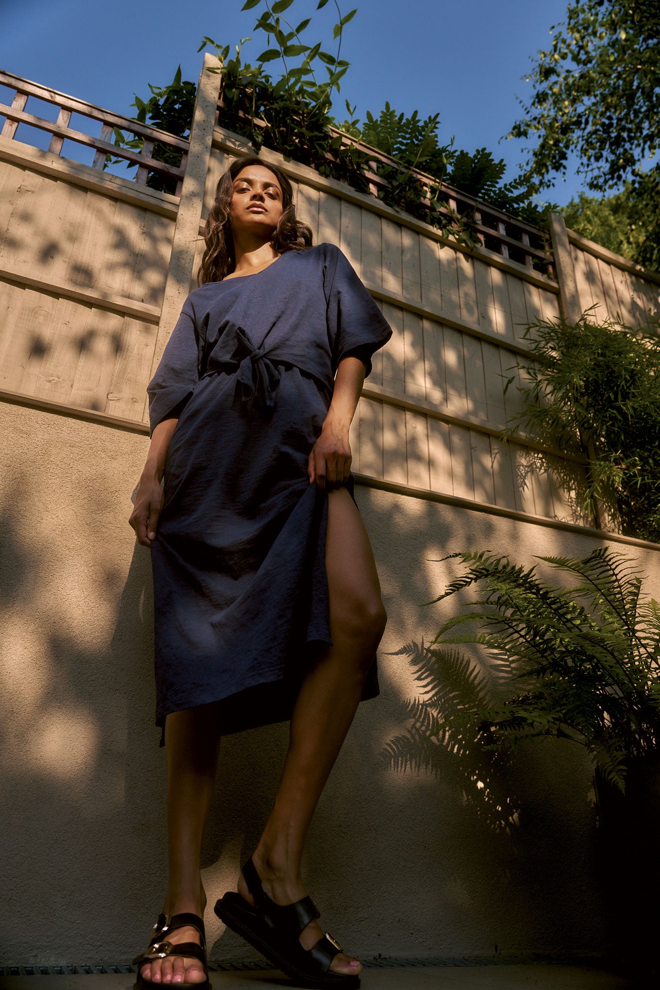 Woman in a navy relaxed dress standing against a wooden fence with plants