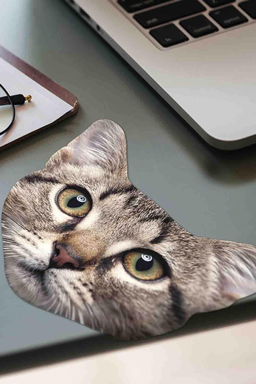 A novelty eyeglass cloth shaped like a tabby cat's face, featuring realistic fur stripes and green eyes, placed on a gray desk next to a laptop and glasses.
