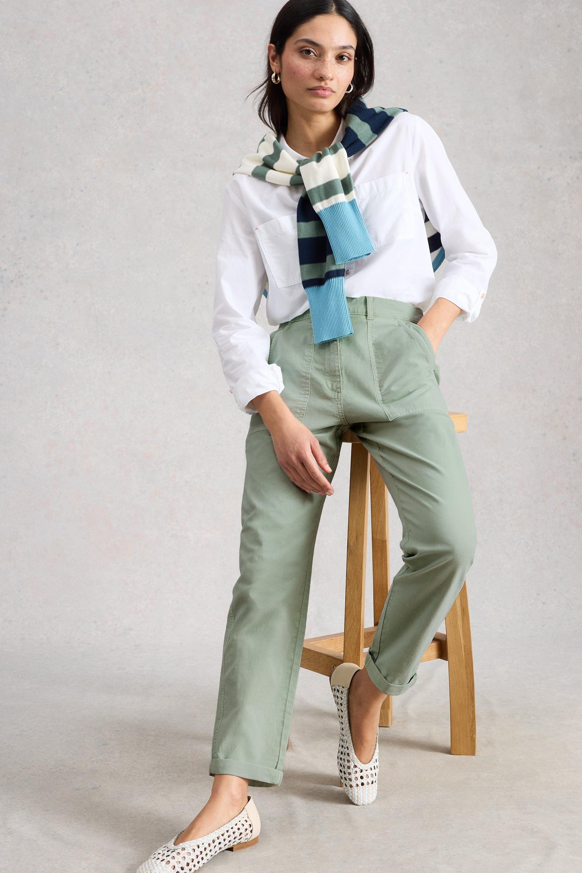 Woman sitting on a stool wearing a white shirt, green organic cotton pants, and a plaid scarf with a blue accent.