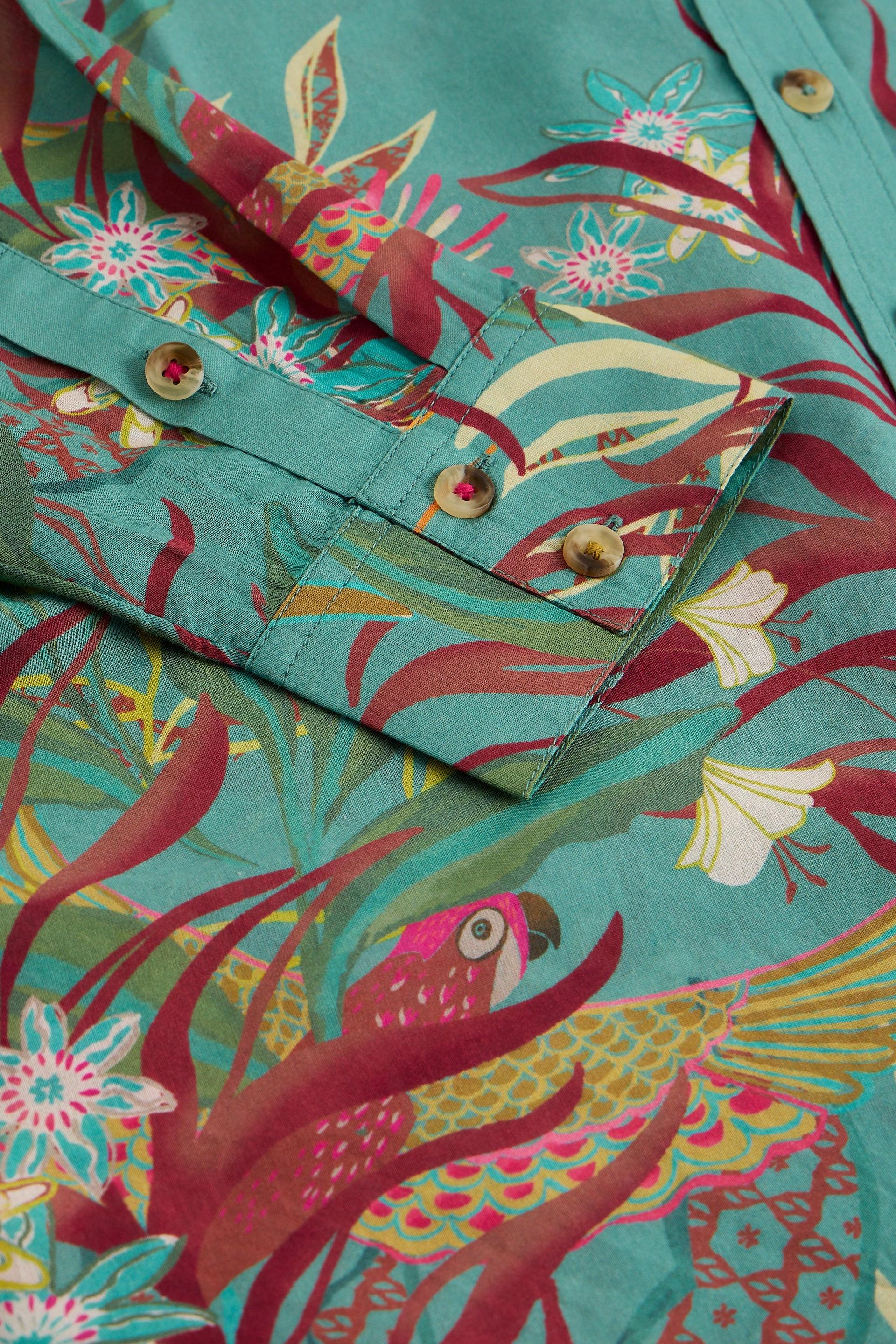 Close-up of a teal organic cotton printed shirt with vibrant floral and parrot patterns and buttoned cuffs.