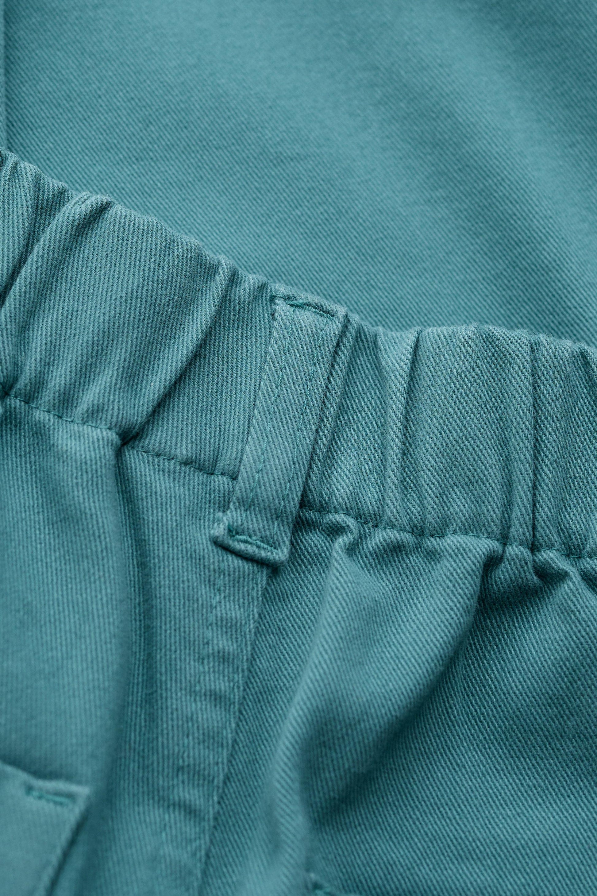 Close-up of a teal fabric garment showing an elasticized waistband with stitched belt loops and a pocket edge.