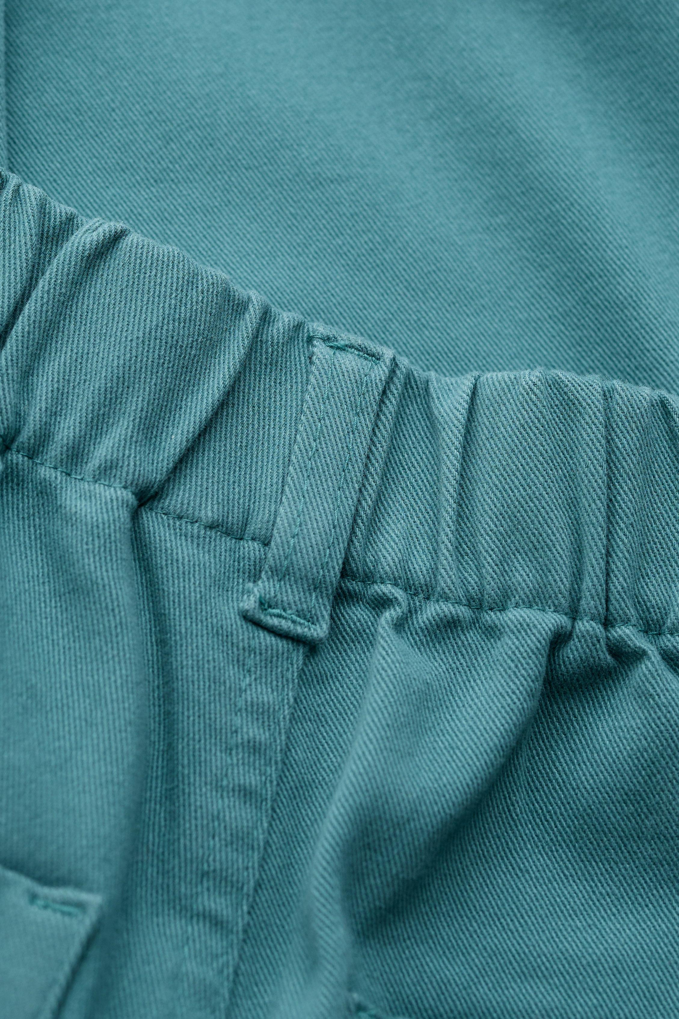 Close-up of a teal fabric garment showing an elasticized waistband with stitched belt loops and a pocket edge.