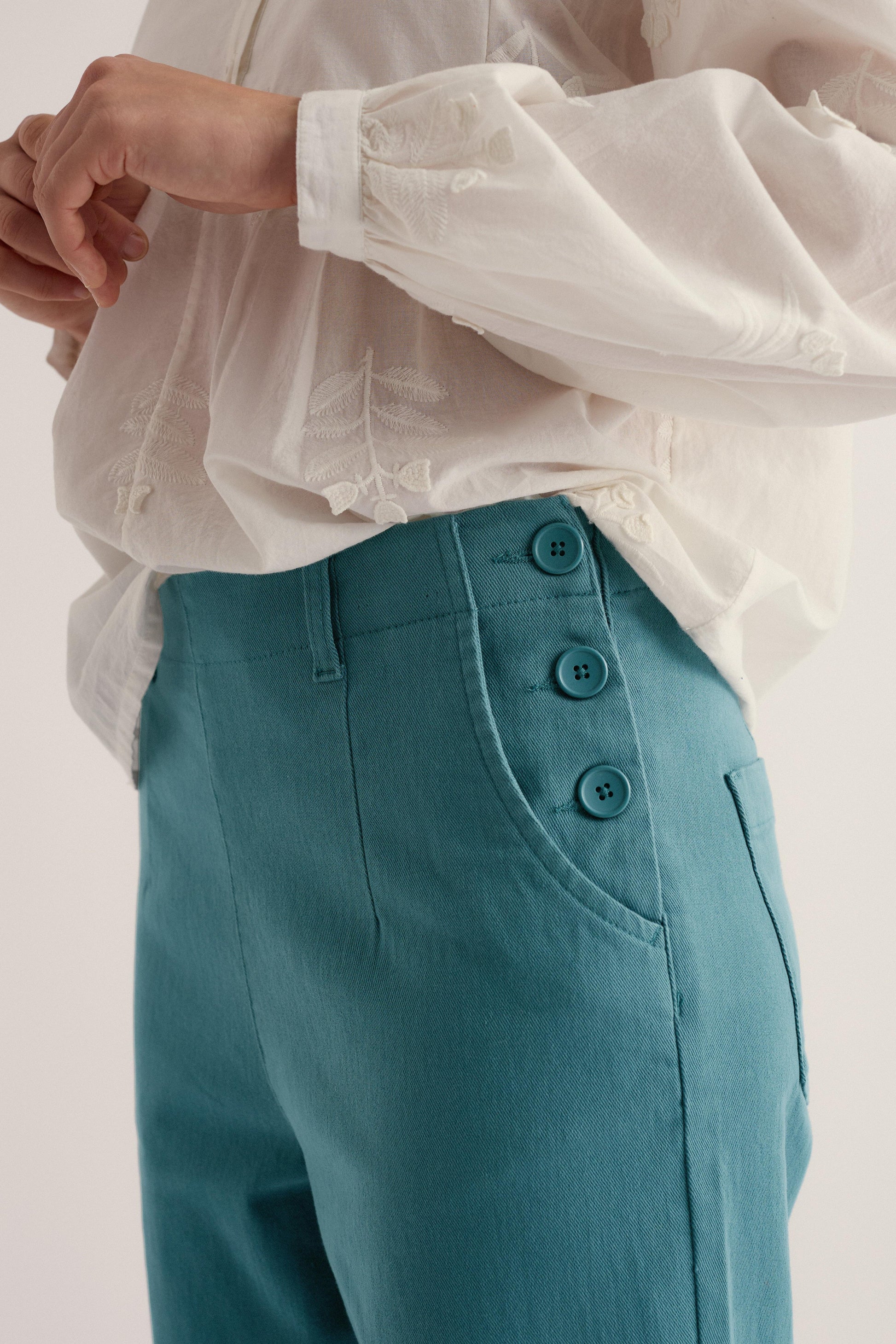 Teal pants with button details worn over a white shirt on a light background