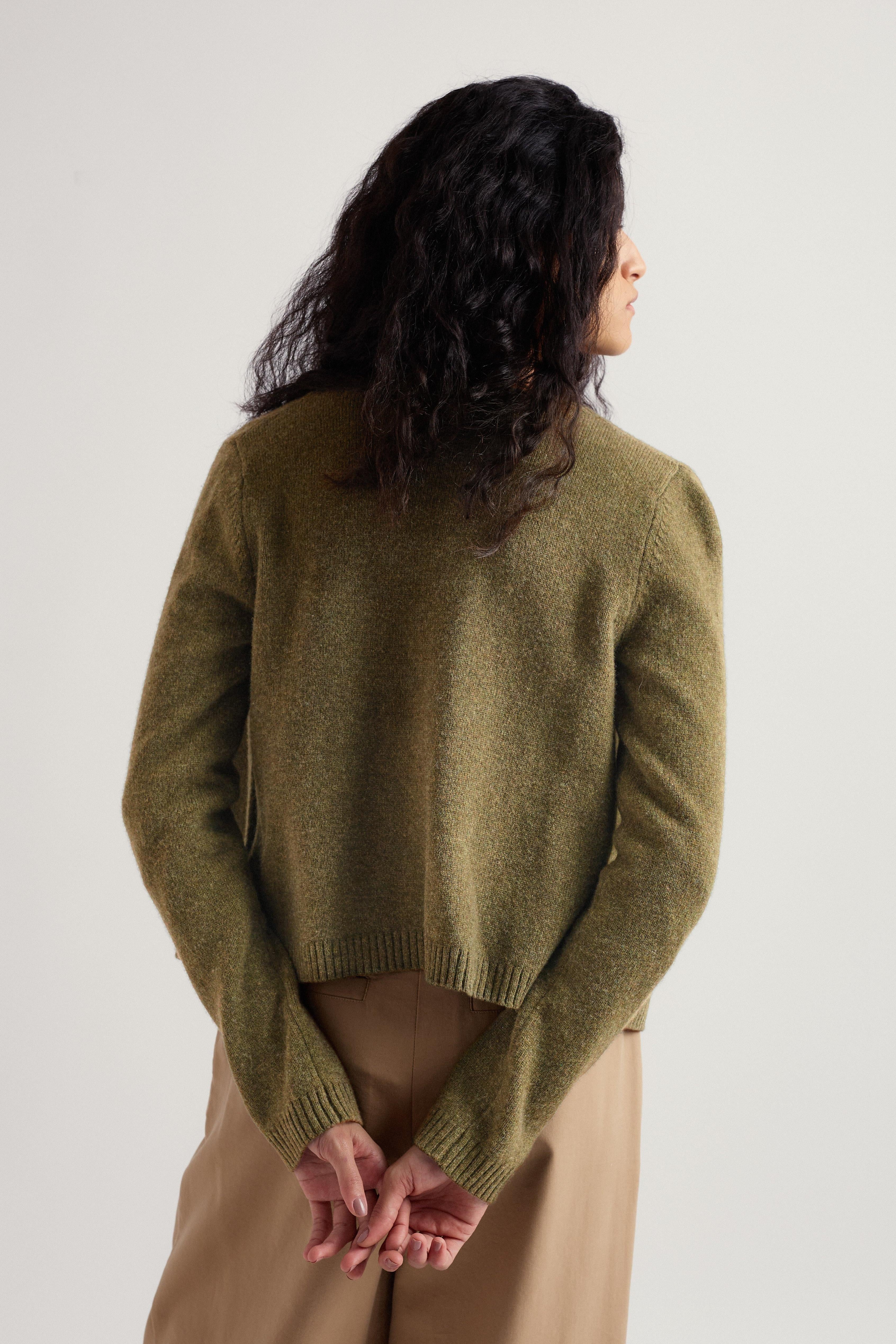 Person wearing a green merino wool cardigan and beige pants against a white background