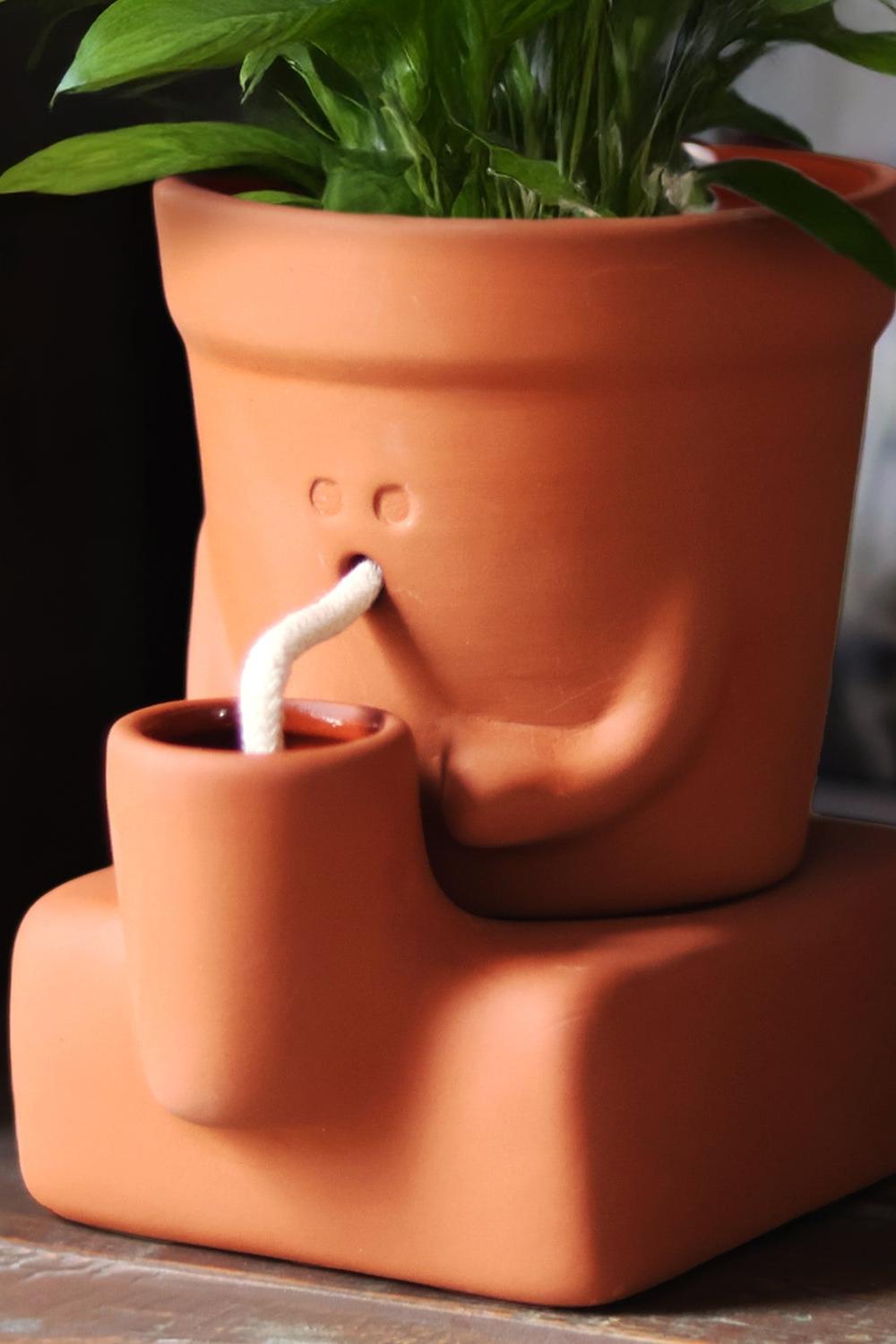 A terracotta-colored ceramic planter shaped like a face, with a small side cup and a white curved spout, holding green leafy stems against a dark indoor background.