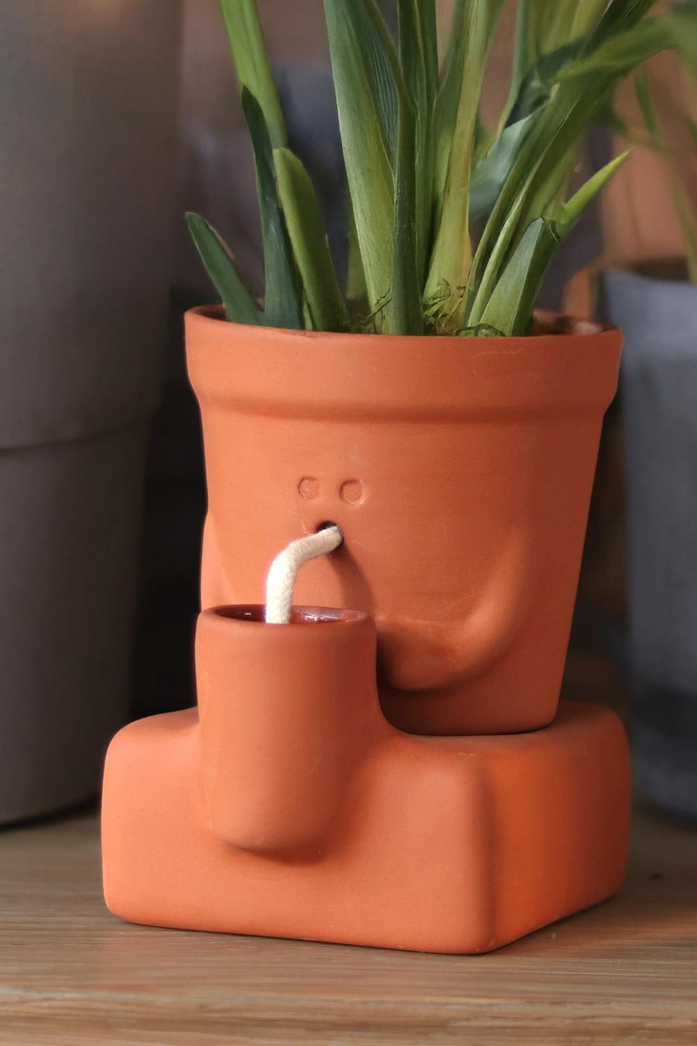 An orange terracotta planter with a built-in mug and a small white cord, shown on a wooden surface with green plants in the background.