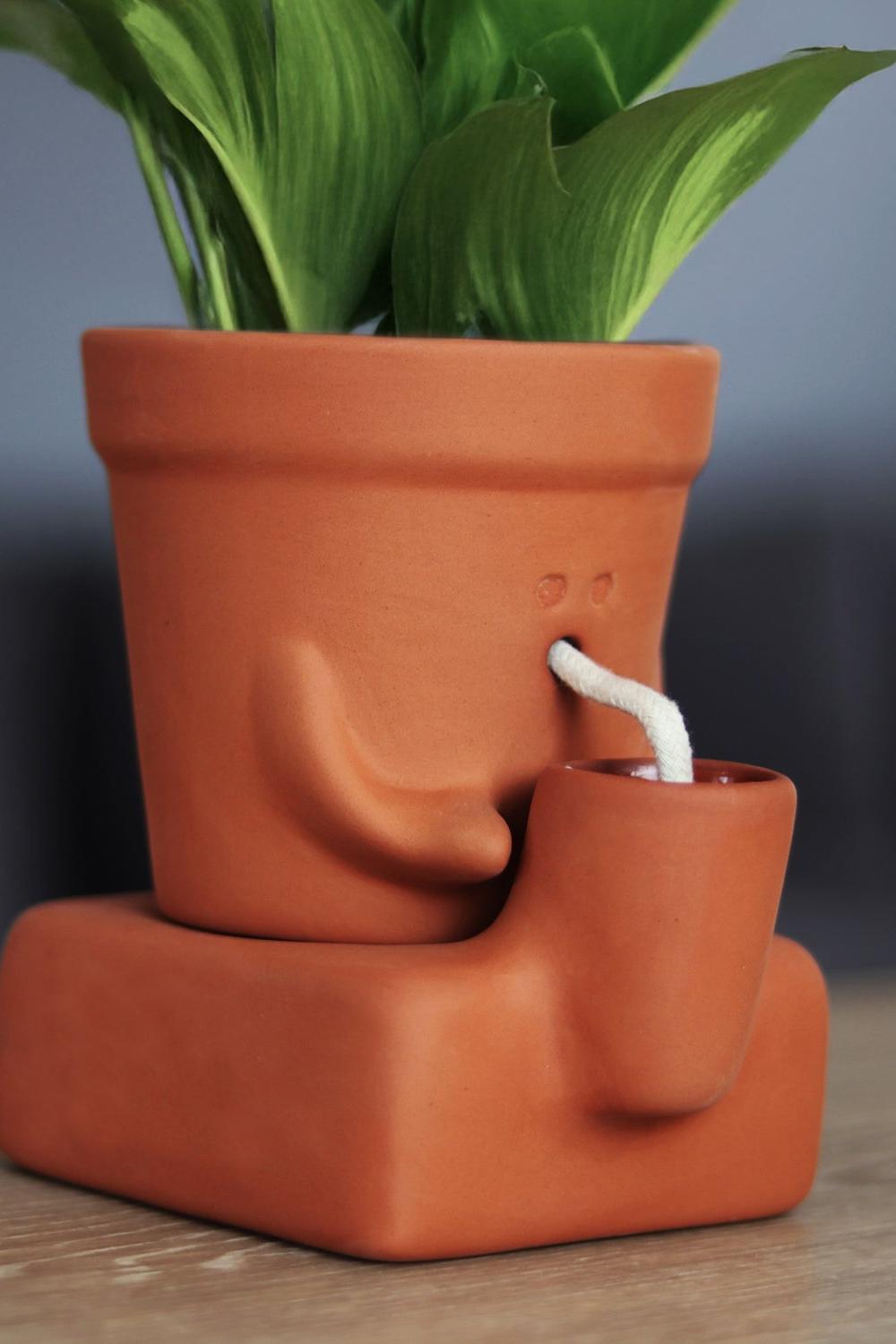 A terracotta-colored ceramic planter shaped like a face with a small protruding cup and white wick, holding green plant leaves, set on a wooden surface with a blurred dark background.