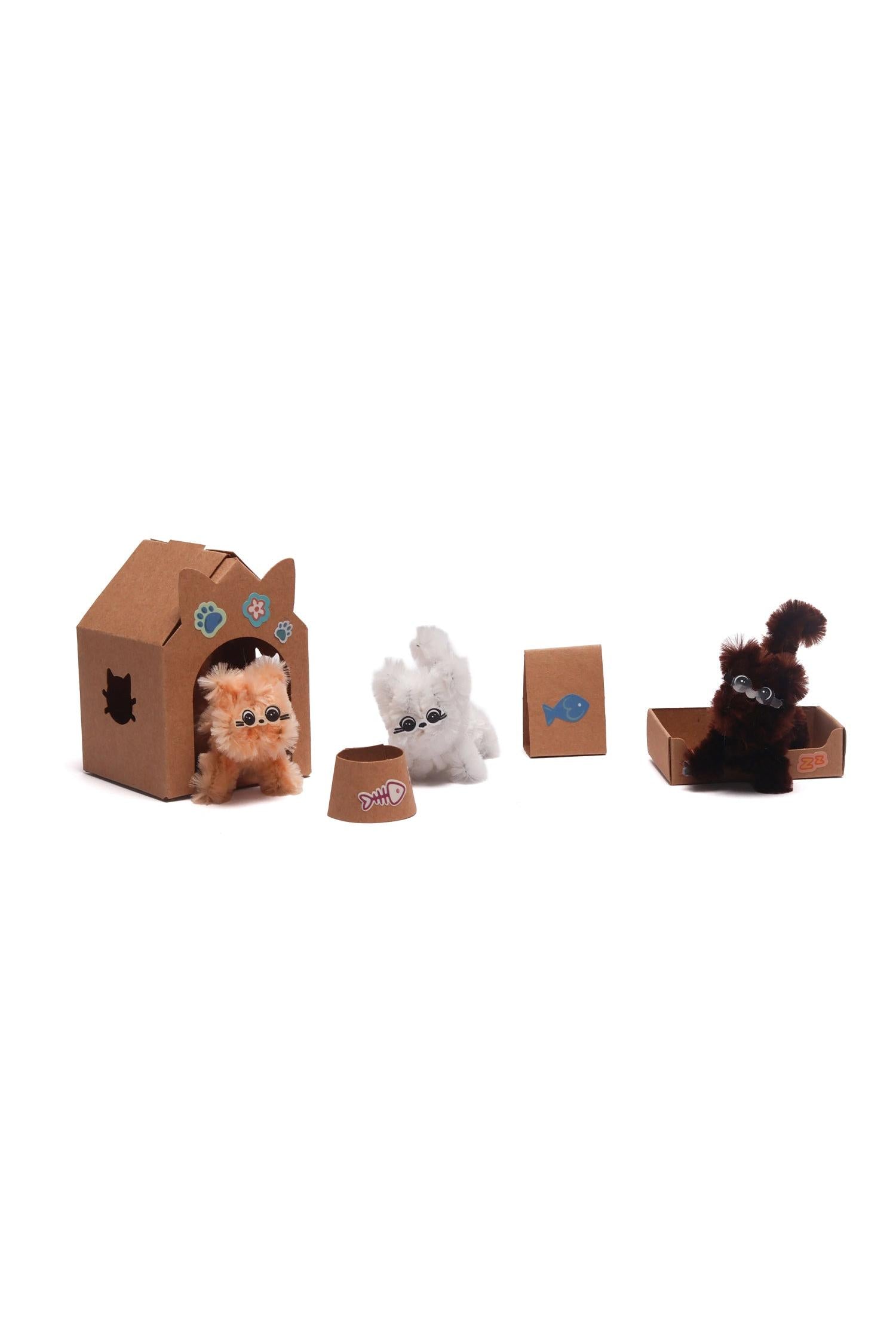 Three small fluffy toy cats in orange, white, and brown with a cardboard cat house, bed, and food bowl.
