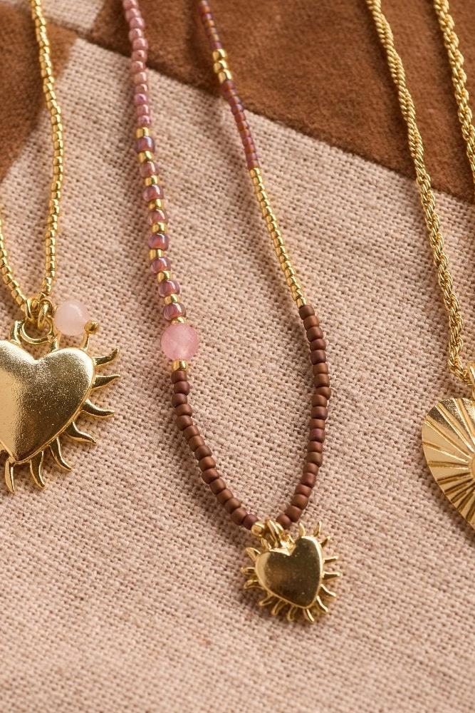 Three gold heart necklaces with rose quartz and gemstone details arranged on a textured checkered fabric background.