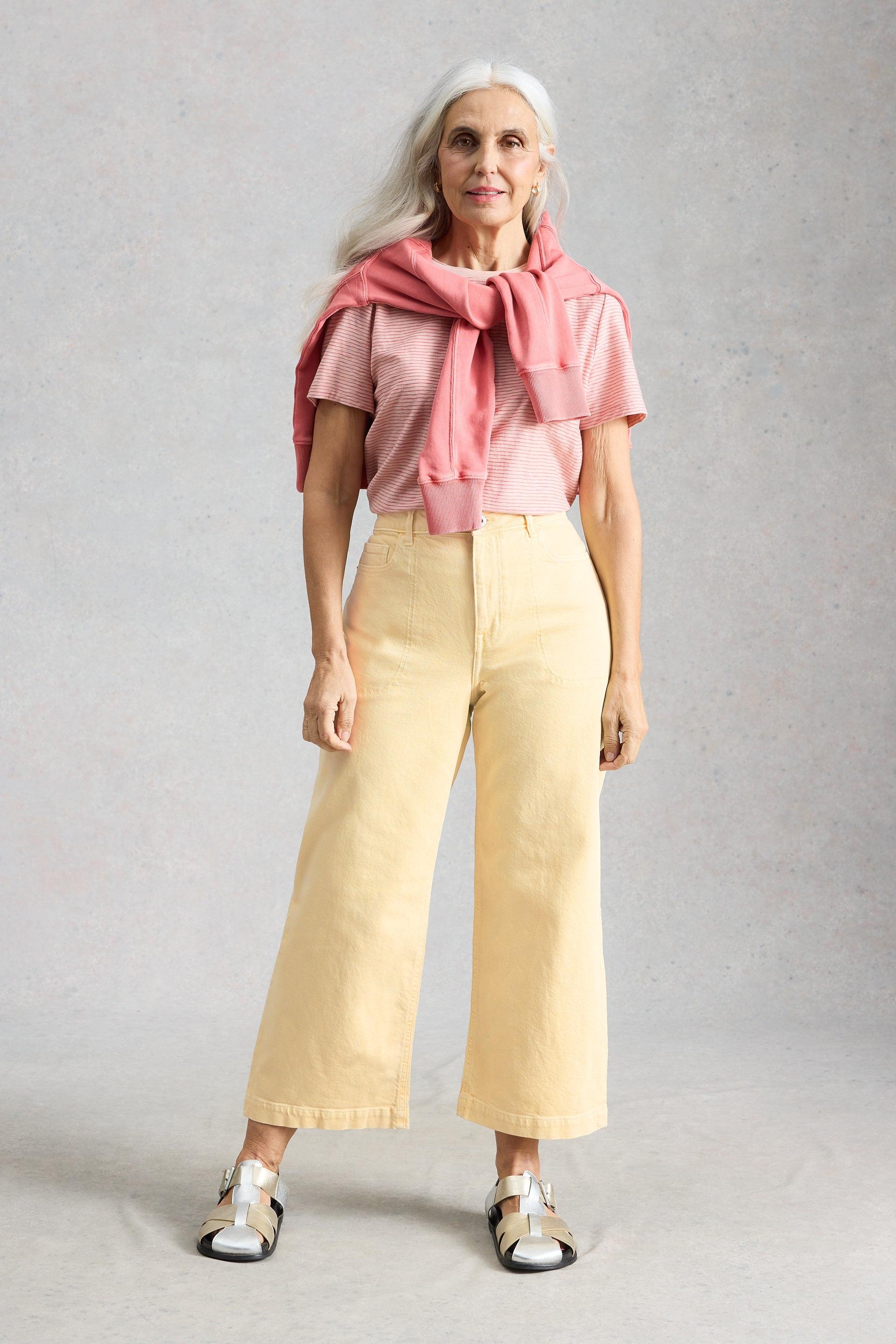 Woman wearing a pink t-shirt and yellow mid-rise jeans with a pink sweater around shoulders on a gray background